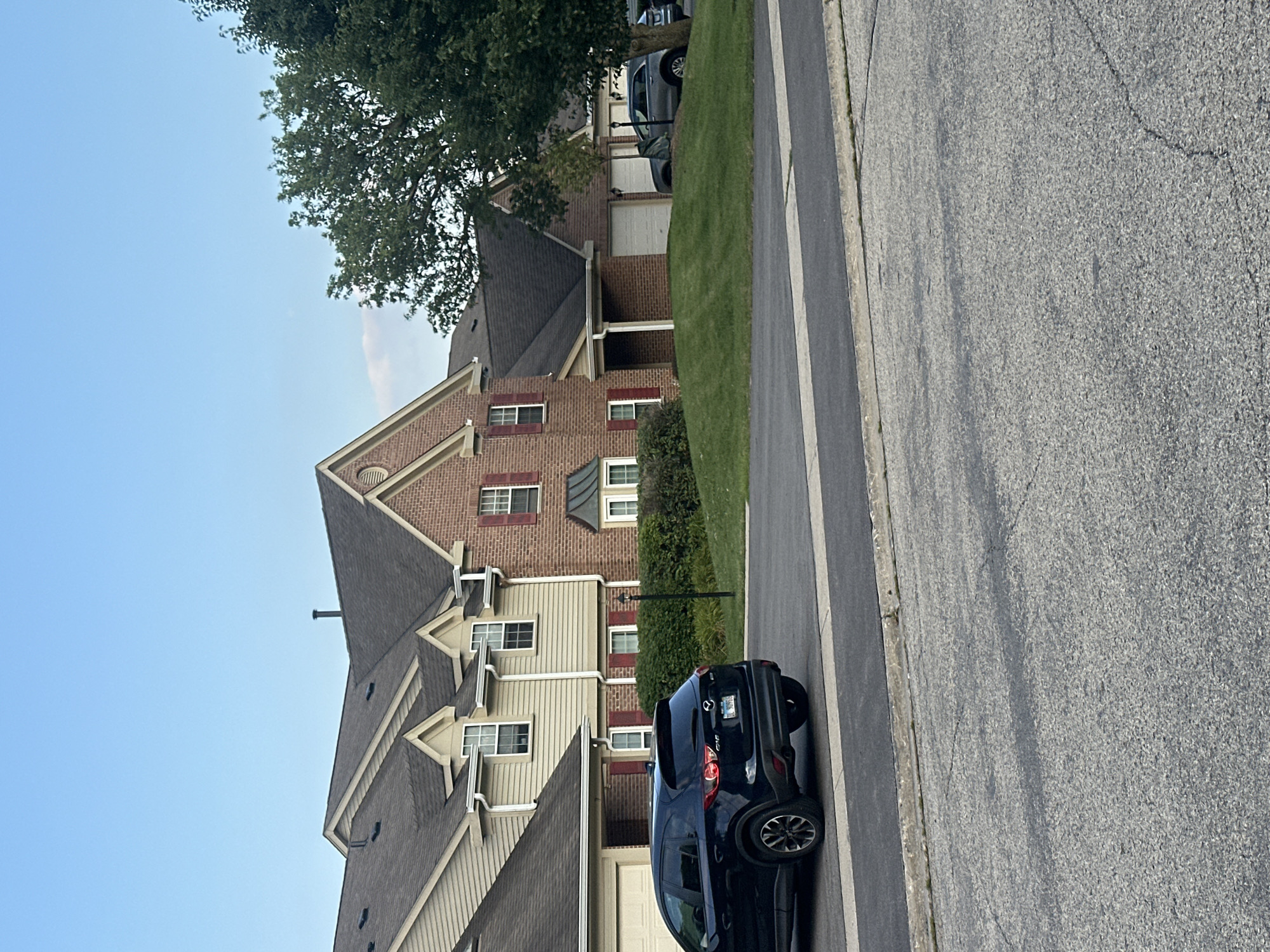 In Schaumburg, Illinois, this image captures a street view in front of residential townhouses. The foreground shows a paved road with a distinct curb and a strip of grass. A dark blue SUV is parked on the right side of the road, angled away from the camera. Beyond the road, a manicured lawn slopes upwards towards the townhouses. These multi-story residences feature a mix of light-colored siding and red brick. Red shutters are visible on some of the windows. A large green tree with dense foliage stands to the right of the townhouses, partially obscuring the sky. The sky is clear and light blue, indicating daytime. There are no people visible in the scene.