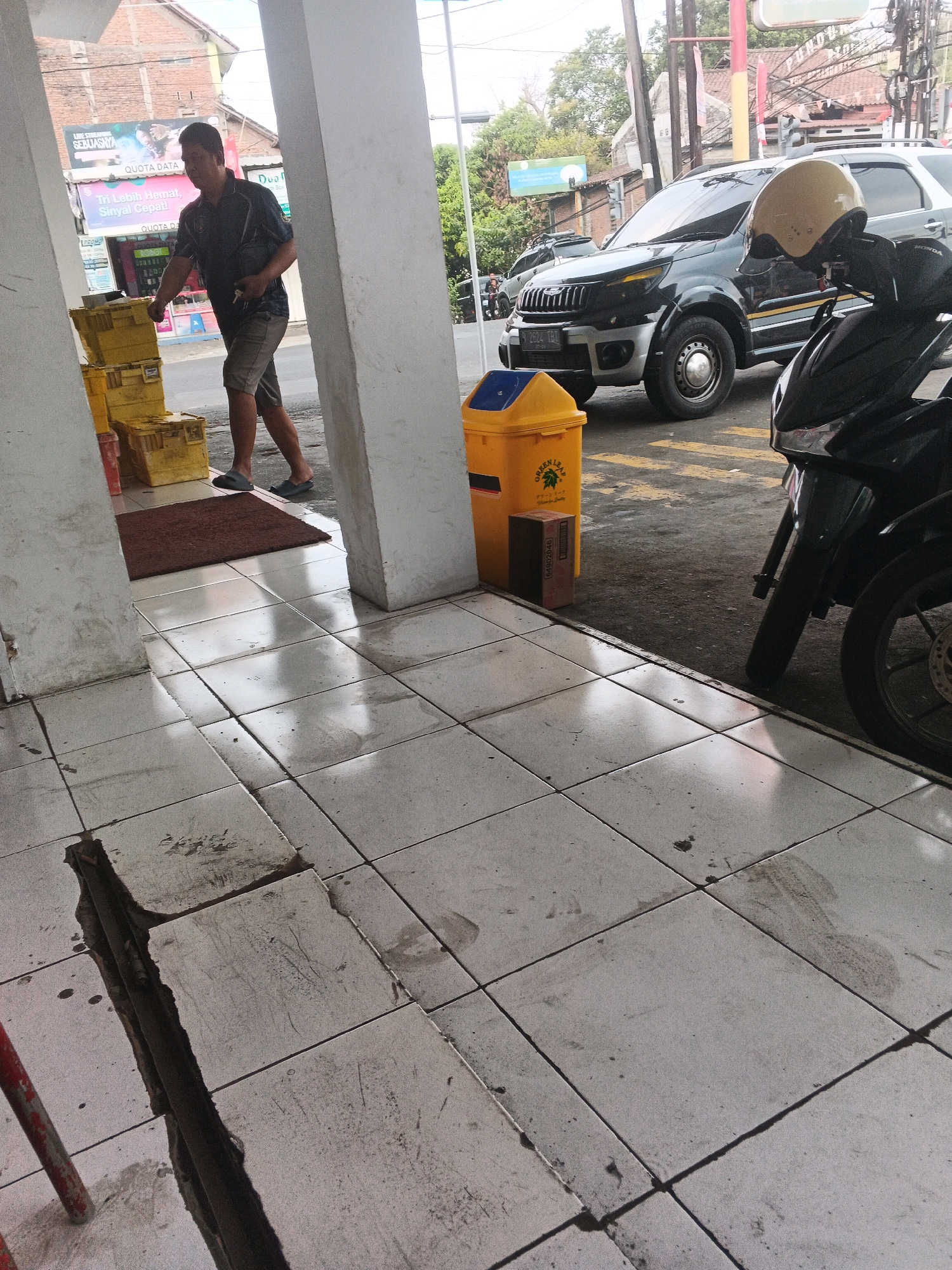 A man in a dark short-sleeved shirt and shorts walks past a stack of yellow plastic crates on the tiled floor in front of a building. He is wearing dark slippers. To his right, a yellow trash can stands next to a building column. A black motorcycle with a yellow helmet on its handlebars is parked to the right of the trash can, partially obscuring a black SUV. The scene appears to be a street in the Genuk Barat area of Indonesia, with buildings and power lines visible in the background. The tiled ground shows signs of wear and some discoloration.