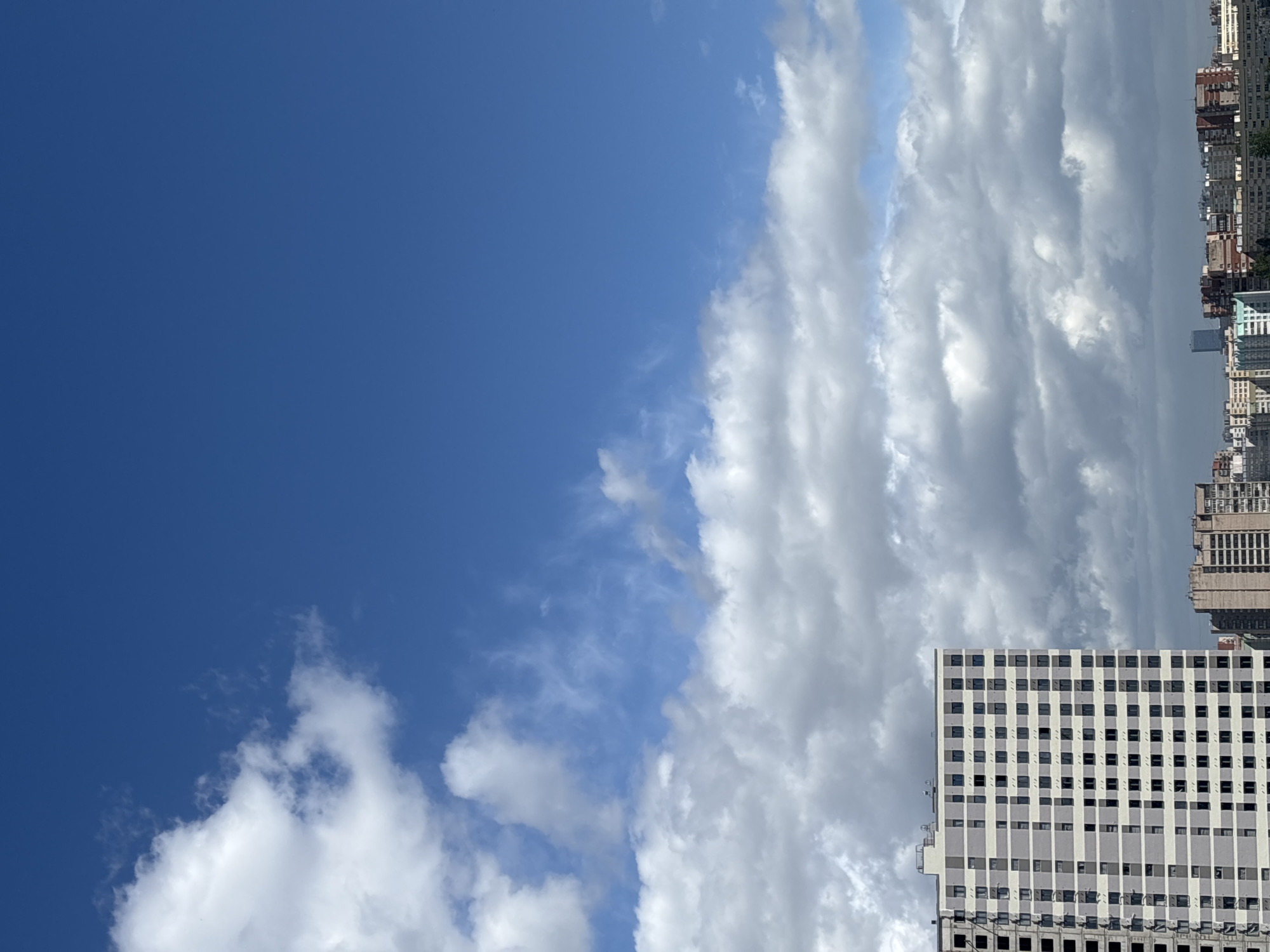 A shot of the upper portion of a building with a grid of windows is positioned in the lower right corner of the image. The building's exterior is in shades of white and gray. Above and to the left of the building, there's a dense arrangement of clouds. The sky on the left side is a clear, deep blue. In the far background, a series of buildings line the horizon. The scene appears to be in an urban environment, with a focus on architecture and the sky.