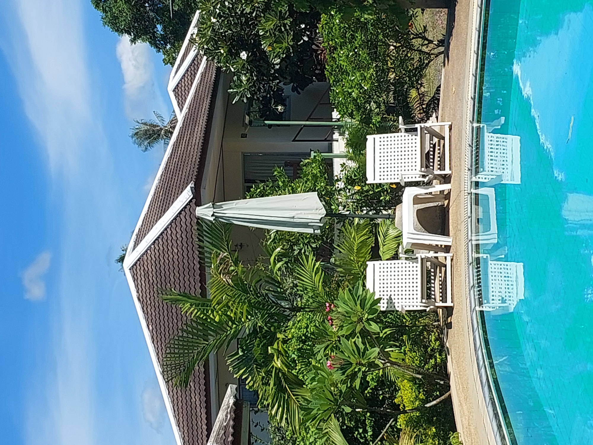 In Baan Mae Nam, Thailand, a poolside scene unfolds under a clear blue sky dotted with white clouds. Two white slatted lounge chairs sit on the tan-colored pool deck, flanking a small white table. An open, light green patio umbrella stands behind the table, its fabric casting a shadow. Lush green foliage, including palm fronds and flowering plants with pink blossoms, surrounds the area, with the tiled roof of a building visible behind the greenery. The bright blue water of the swimming pool occupies the foreground, its surface reflecting the chairs and the sky.