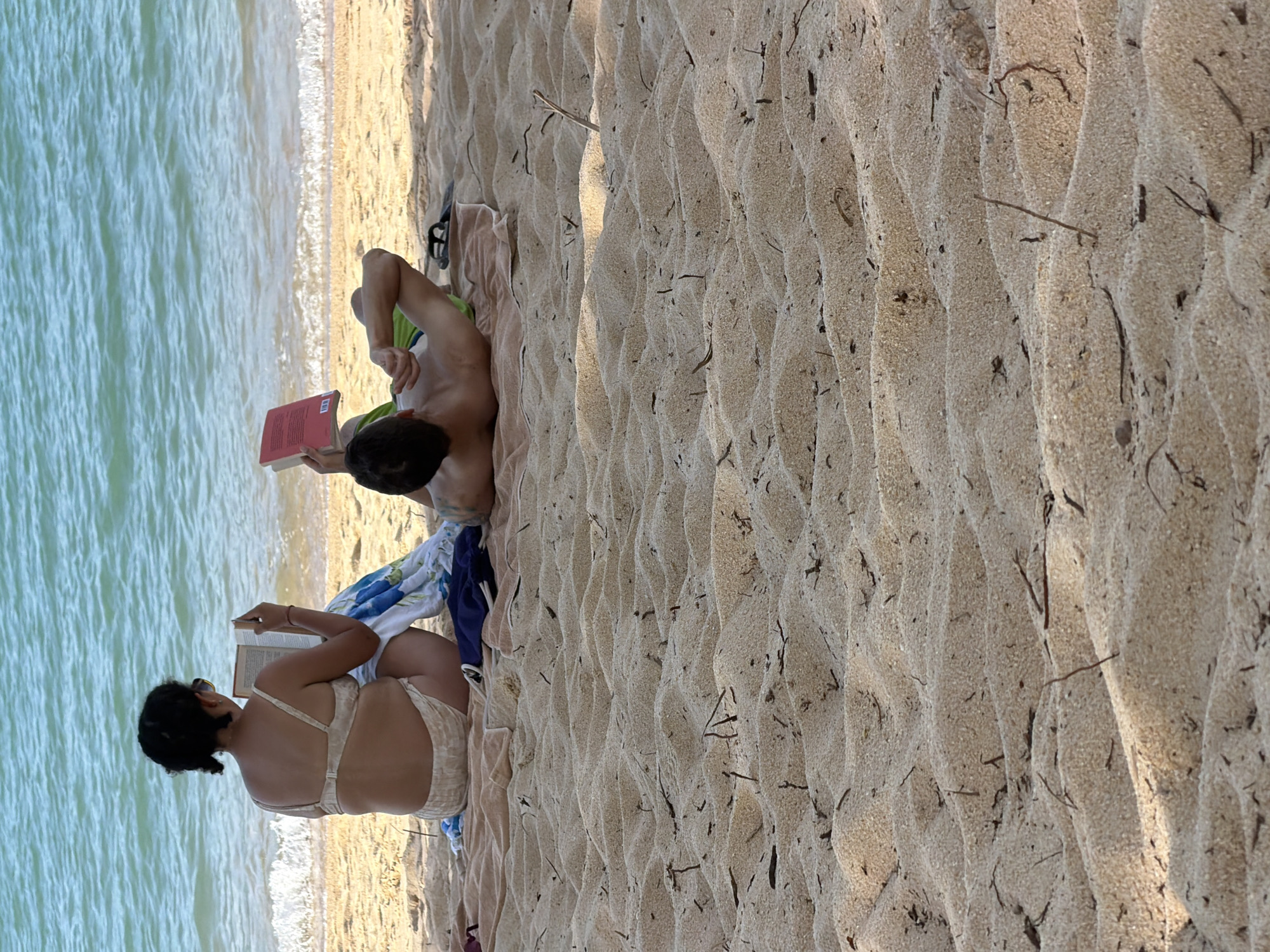 Two individuals are seen relaxing on a sandy beach in Baan Tai, Thailand, near the ocean. In the foreground, the sand is textured with small ripples and scattered pieces of dried seaweed. The middle ground features two towels laid out on the sand. On the left, a person wearing a light-colored bikini is seated and reading a book. On the right, another person is lying on their back on a towel, also reading a book. The ocean is visible in the background with gentle waves breaking on the shore. The sky is not visible in the frame.