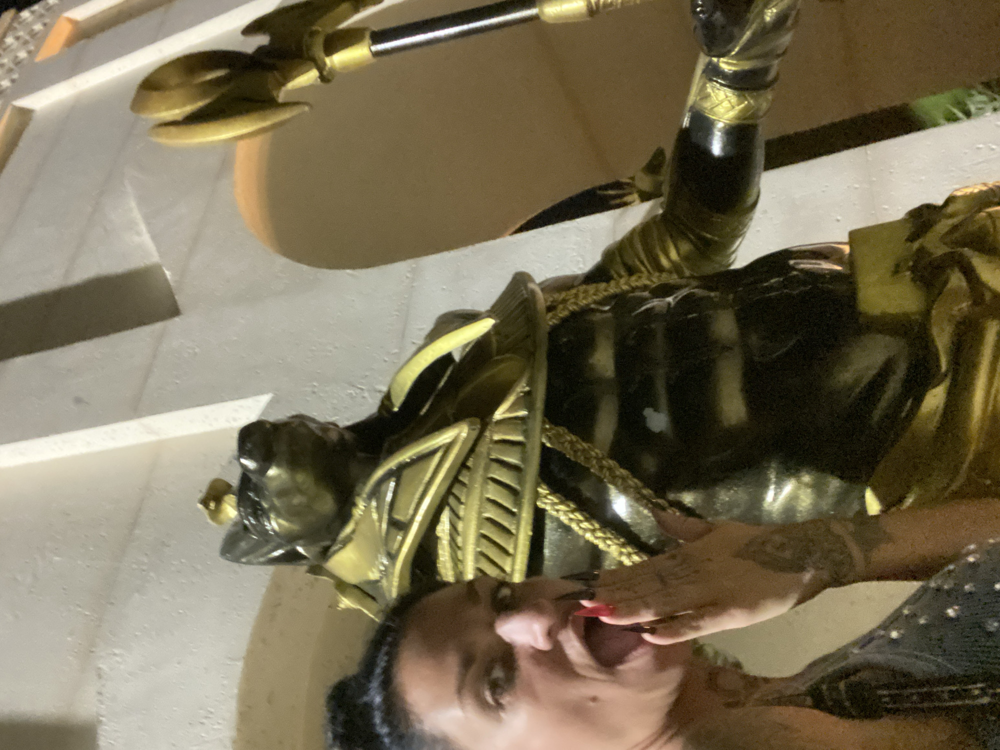 A person is pictured in front of a statue. The statue depicts a figure adorned in gold, black, and bronze armor, holding a scepter. The person is partially visible, with their face and part of their upper body in view, and is holding their hand near their mouth. The environment appears to be an outdoor location, potentially a public space. Based on the visible elements, this image was taken in Mũi Né, Vietnam, at the landmark for the "Poseidon Resort" which has the large "Poseidon" statue in front of it.