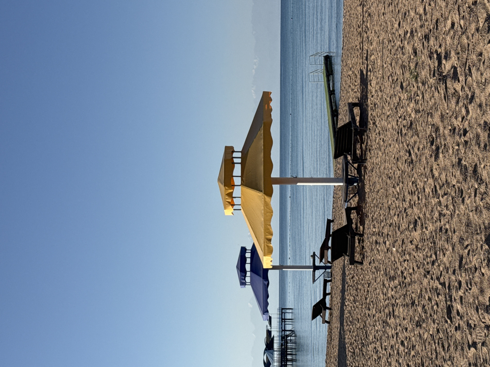 This is a photograph of a beach scene in Sary-Oy, Kyrgyzstan. In the foreground, there is a sandy beach with several empty lounge chairs and umbrellas. The umbrellas are yellow and blue. In the middle ground, there is a calm body of water, likely Lake Issyk-Kul, with a pier extending into it. In the background, there are mountains visible. The sky is clear and blue, indicating a sunny day. The scene appears to be set up for relaxation and leisure.