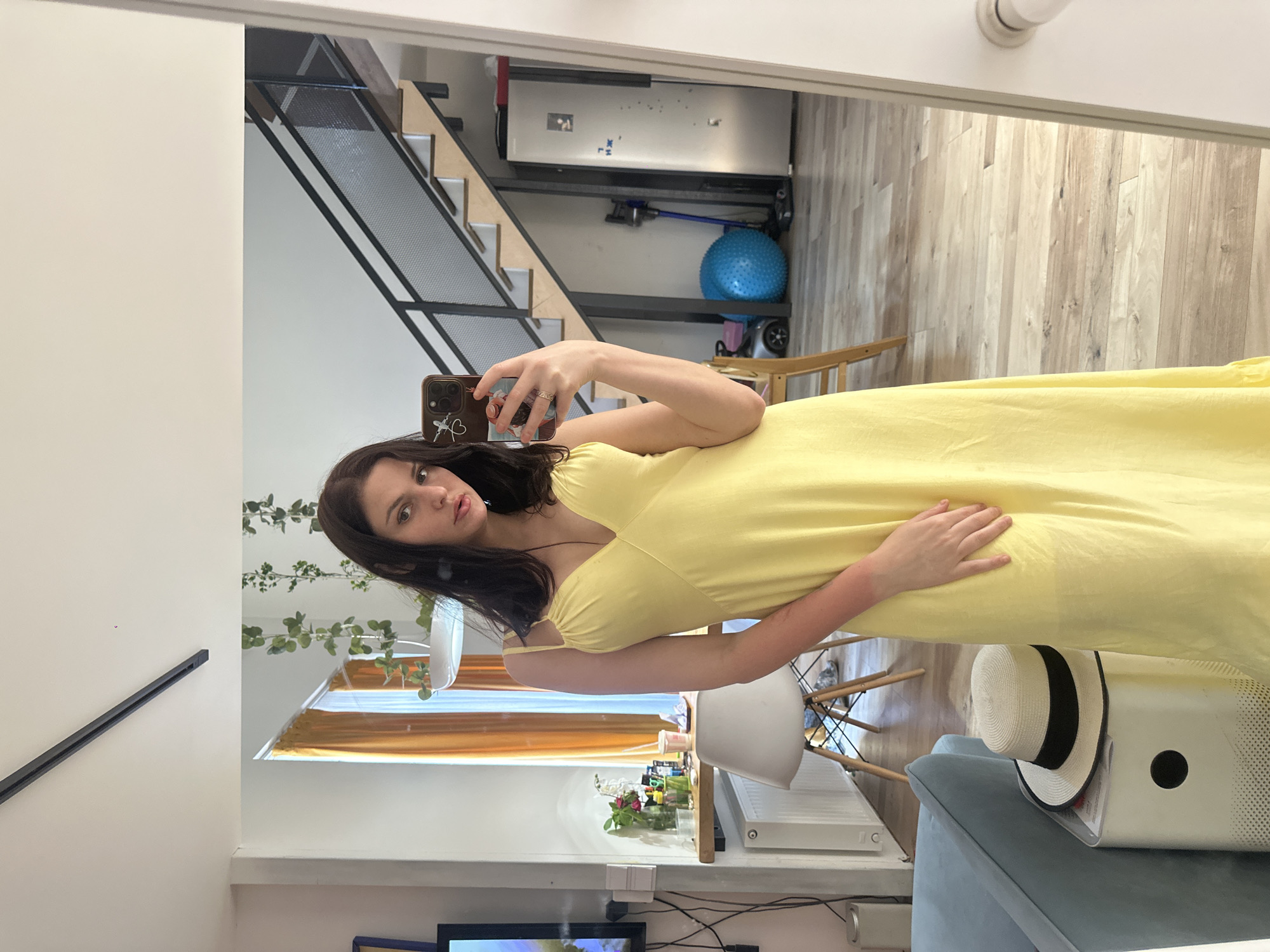 In the provided image, a young woman is taking a mirror selfie in a brightly lit room. She is wearing a flowing yellow dress and has long, dark hair. Her expression is neutral, and she is holding a smartphone in her right hand to capture the reflection.
The background of the reflection shows a modern interior. To the left, a staircase with black metal railings and wooden steps is visible, leading upwards. Above the staircase, shelves are mounted on the wall, holding various items, including a blue exercise ball and what appears to be a vacuum cleaner. The flooring visible in the reflection is light-colored wood.
To the right, the scene includes a white chair, a radiator, and a blue sofa with a white and black striped hat resting on a white object. There are also some plants and decorative items on a shelf. Sunlight streams in from a window, casting a warm glow on the scene. The overall impression is of a home environment during the daytime.