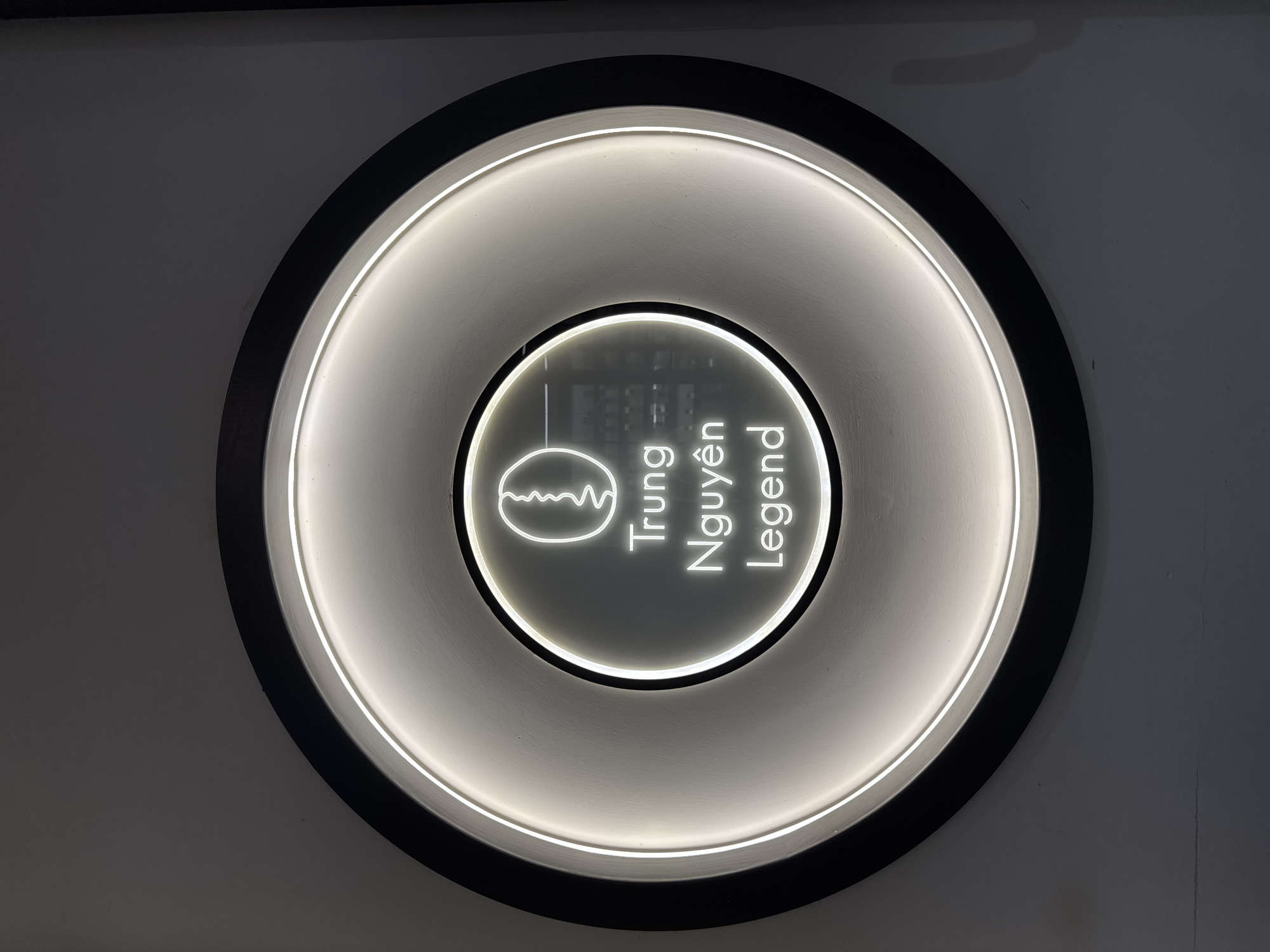 This media file shows a close-up, top-down view of a circular, illuminated sign mounted on a wall. The sign features a black outer rim and a lighter, textured inner circle with a bright, white neon-like light tracing the edges of concentric rings within it.
At the center of the sign, within the innermost illuminated circle, is a stylized outline of a coffee bean. To the right of the coffee bean, stacked vertically in white neon text, are the words "Trung Nguyên Legend".
The lighting suggests the sign is indoors or in a dimly lit area, possibly at night, designed to be eye-catching. The overall aesthetic is modern and minimalist, with a focus on the brand's name and logo. There are no people or specific environmental details visible that would indicate the time of day or weather conditions, other than the artificial lighting of the sign. The location context of Nha Trang Ward, Vietnam, points to the origin of the brand, which is a well-known Vietnamese coffee producer.