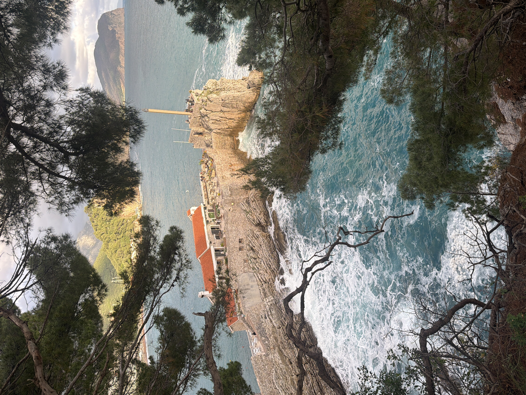 The image, rotated counter-clockwise, shows a high-angle view of the coastline near Petrovac centar, Montenegro. The Adriatic Sea, displaying turbulent white waves, fills a large portion of the scene, framed by trees in the foreground. A long, narrow peninsula extends into the sea, topped by a stone building with a red-tiled roof. A stone wall follows the edge of the peninsula. A slender, tall pole stands on the tip of the peninsula. A mountainous coastline is visible in the background.