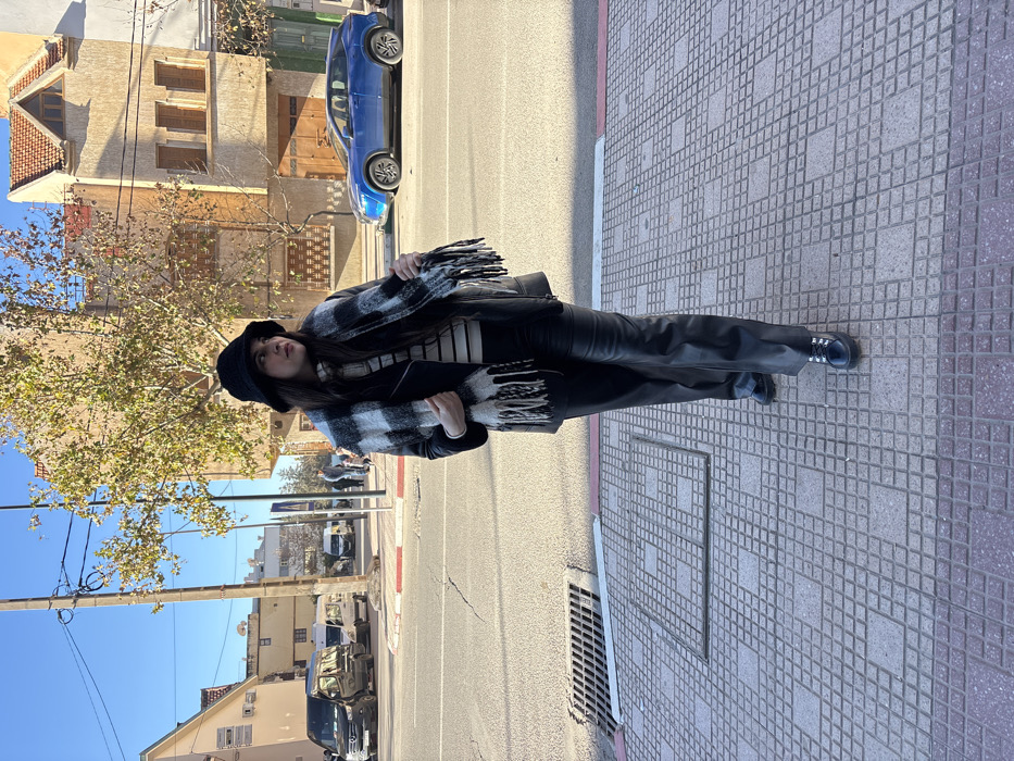 The image depicts a female subject standing on a tiled sidewalk in the city of Azrou, Morocco, under a clear blue sky. She is positioned slightly right of center, facing towards the right side of the frame. Her attire consists of a black bucket hat, a black leather-like jacket worn over a white and black horizontal striped turtleneck sweater, black leather-like trousers, and black patent leather shoes with silver metallic eyelets. A black and white plaid scarf with fringed ends is draped over her shoulders, with her left hand appearing to adjust or hold one of the fringed ends.
The sidewalk beneath her is composed of light-toned, square tiles arranged in a grid pattern. A dark metal drainage grate is visible on the left side of the sidewalk. Adjacent to the sidewalk is an asphalt road, showing signs of wear and cracking, bordered by a white line and a red painted curb line.
In the background, on the right side of the street, a blue compact SUV is parked, facing left. On the left side of the street, further down, a black van and several white vans are visible. The dominant building in the immediate background on the right is a two-story structure with a light brown or tan stucco-like facade, featuring multiple windows (some with closed wooden shutters) and a prominent reddish-orange tiled gable roof. A deciduous tree with sparse, yellowish-brown foliage stands between the street and this building. Additional multi-story buildings are visible further in the background on the left. The scene is illuminated by bright daylight.