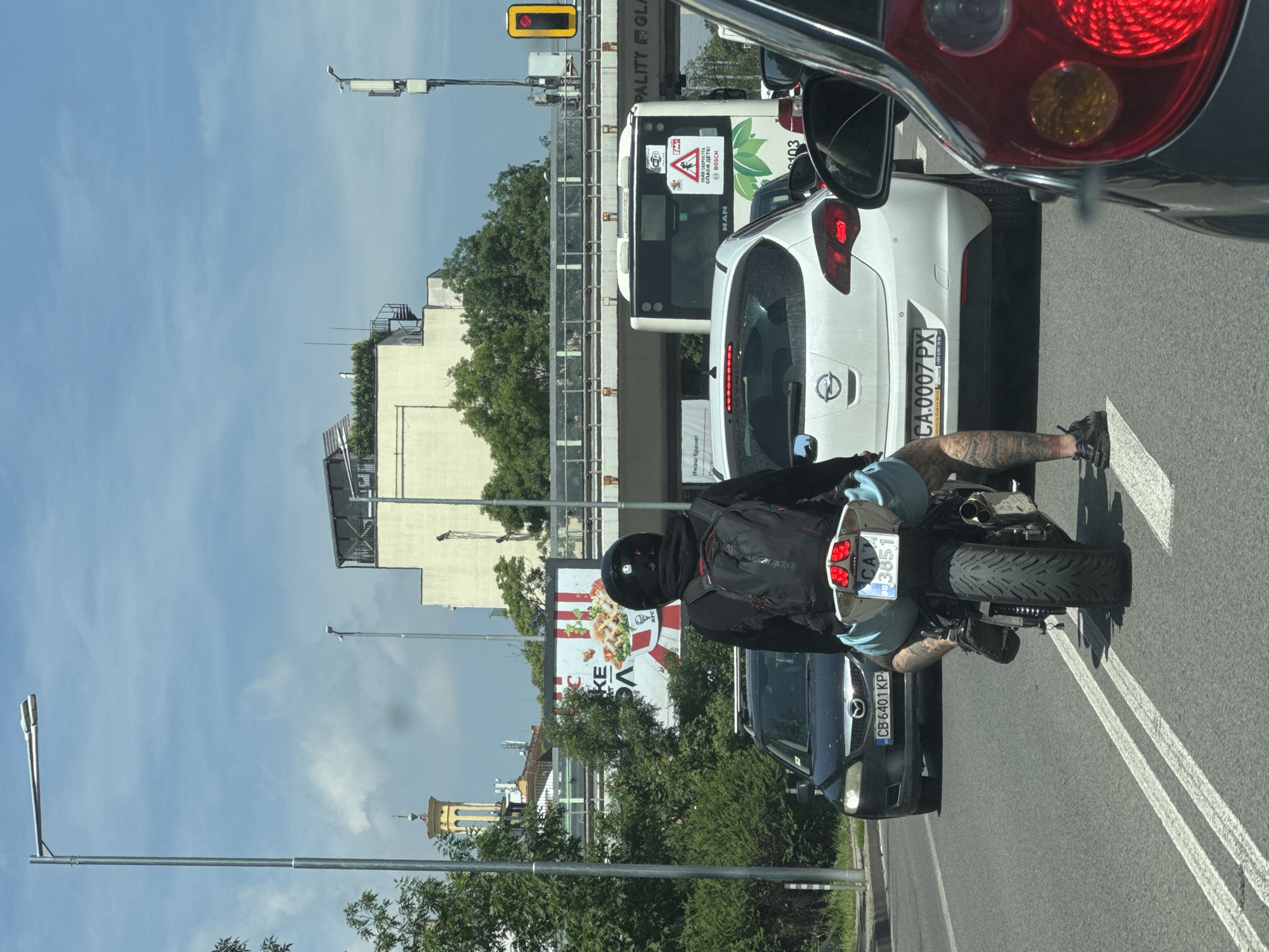 The image depicts a daytime traffic scene in Sofia, Bulgaria, captured from inside a vehicle. The sky is clear blue with minimal clouds.
In the immediate foreground, a person on a motorcycle is waiting in traffic. The motorcyclist is wearing a black helmet, a black jacket, light blue shorts, and a black backpack. Notable tattoos are visible on their legs. The motorcycle's license plate is "CA 3851".
Ahead of the motorcycle, a white Opel Astra car is visible, bearing the license plate "CA.0007 PX". Behind the Opel and partially obscured is a white bus, identified by the "MAN" logo. The bus displays a prominent warning sign with Cyrillic text "УБИЙ СКОРОСТТА СПАСИ ДЕТЕ!" (Kill the speed, save a child!), along with "BOSCH", "WiFi", and the number "103". Another dark car with the license plate "CB 6401 KP" is partially visible behind the motorcyclist.
The background shows urban architecture, including various buildings, trees, and streetlights. A large billboard with a KFC advertisement is visible, displaying "KFC" and partially obscured Cyrillic text "КЕ...А". Another text "Имаш време!" (You have time!) is also present on a nearby sign. In the distance, a distinctive yellow-domed church spire can be seen against the sky. A red traffic light is visible above the street, suggesting the vehicles are stopped. The "CA" and "CB" prefixes on the license plates are specific to Bulgaria, with "CA" indicating Sofia.