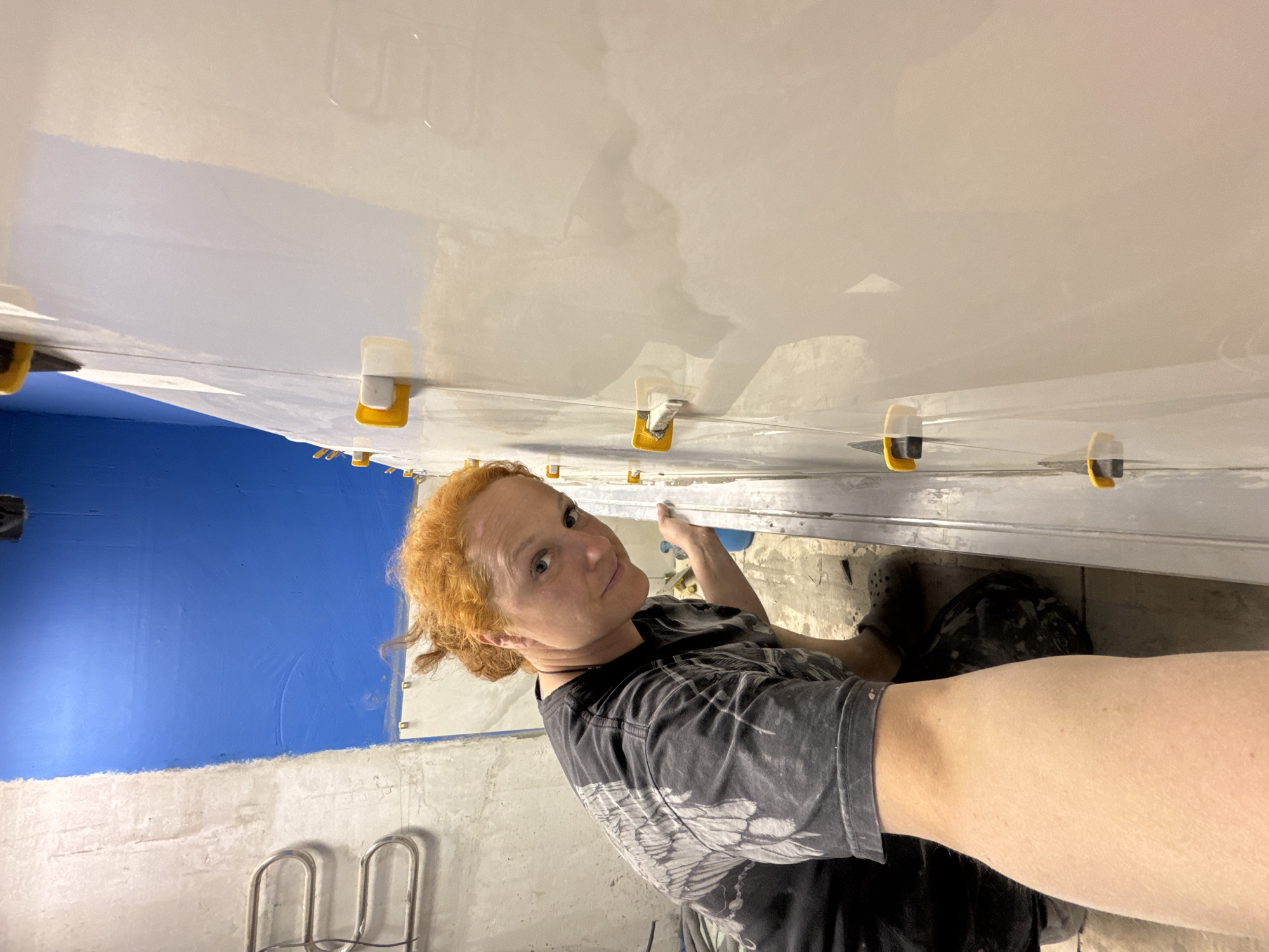A woman with red hair is positioned in front of a wall covered with large, light-colored tiles. Yellow and white leveling clips are used to install the tiles. Behind the woman, a portion of the wall is painted blue. A ladder and what appears to be plumbing are visible on the left side of the frame. The location is a room under construction, possibly in the city of Chaikovsky, Russia. The woman is wearing a dark-colored t-shirt. She appears to be looking directly at the camera, possibly taking a selfie or having someone take the photo while she is working on tiling.