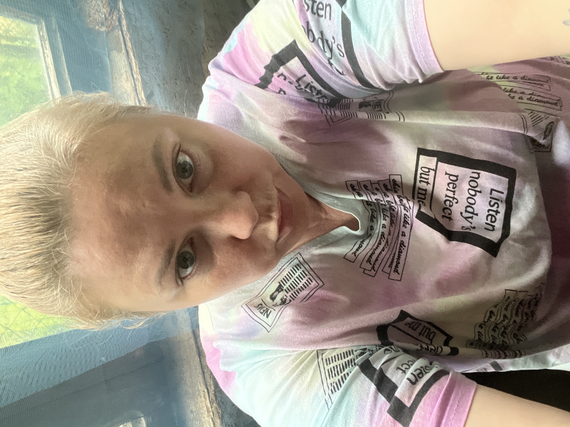 The image is a close-up, angled selfie of a woman with fair skin and blonde hair pulled back. She is wearing a t-shirt with a pastel tie-dye pattern and various text boxes printed on it, including phrases like "Listen nobody's perfect but me" and "Be like a diamond." The text boxes have a news-like design. The background shows a window with a bluish or translucent curtain or netting. The setting is nondescript and does not provide enough information to determine if it is in Vesyoloye, Russia.