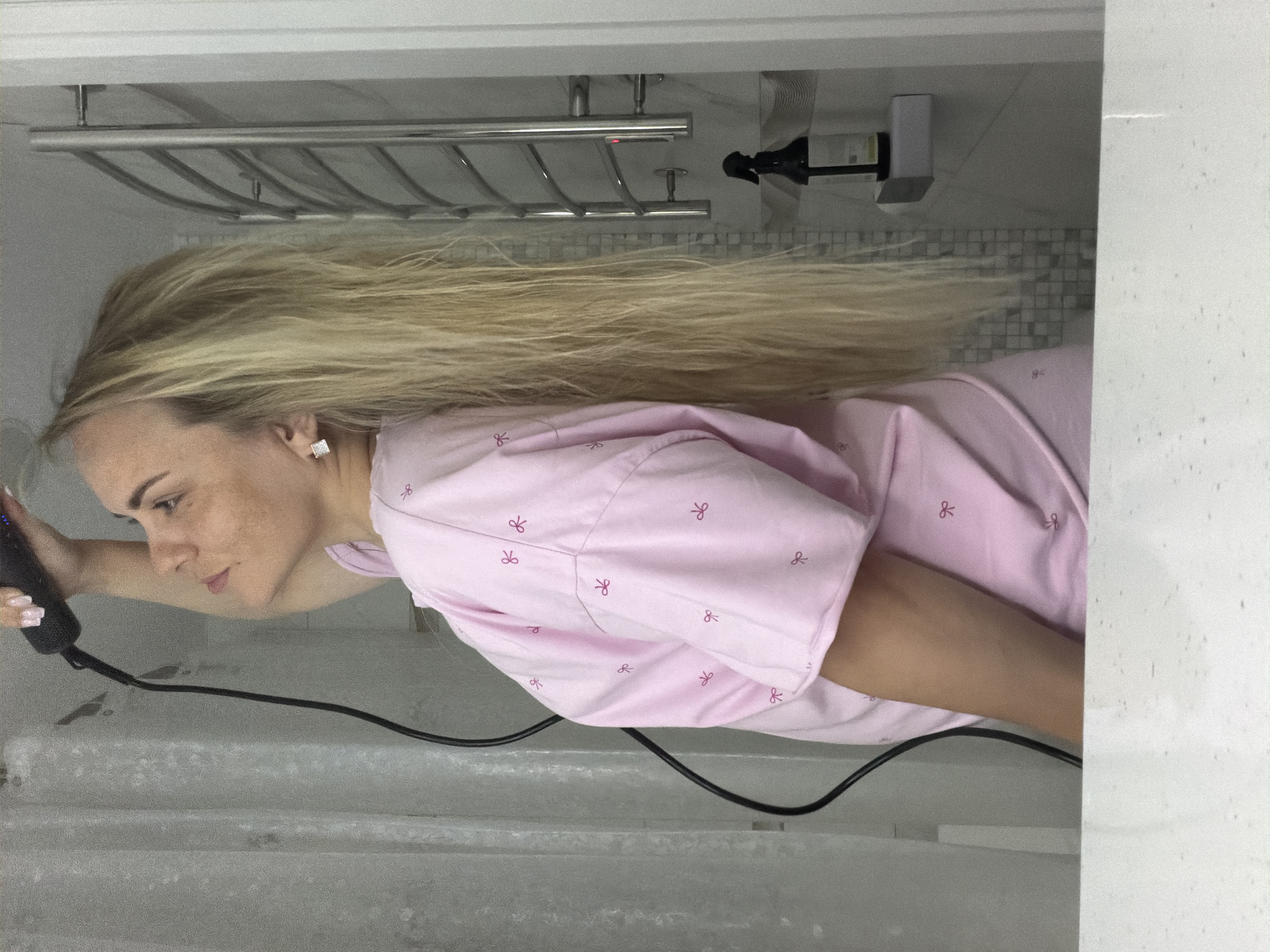 A woman with long, straight, blonde hair is depicted in a side profile, looking downwards and to her right, in an indoor setting, likely a bathroom in Votkinsk, Russia. She has fair skin with visible freckles and wears a light pink garment, possibly a nightgown or shirt, patterned with small, repeating dark pink bows. A sparkling square earring adorns her visible right ear.
She holds a black hair tool, possibly a hair dryer or straightener, in her left hand, which is raised. A black power cord extends from the tool, dropping towards the foreground, which features a light grey, textured surface, possibly a tub edge or counter.
The background shows typical bathroom elements. A chrome-finished multi-tiered towel rack is mounted on a light grey wall above her head. Further to the right, a dark liquid dispenser with a pump top rests on a white surface, set against a section of wall with small, light grey square tiles. The lighting suggests an indoor environment, and the woman's expression is focused as she appears to be styling her hair. The patterns on her clothing, which resemble small bows, were the only elements misinterpreted as text by the OCR.