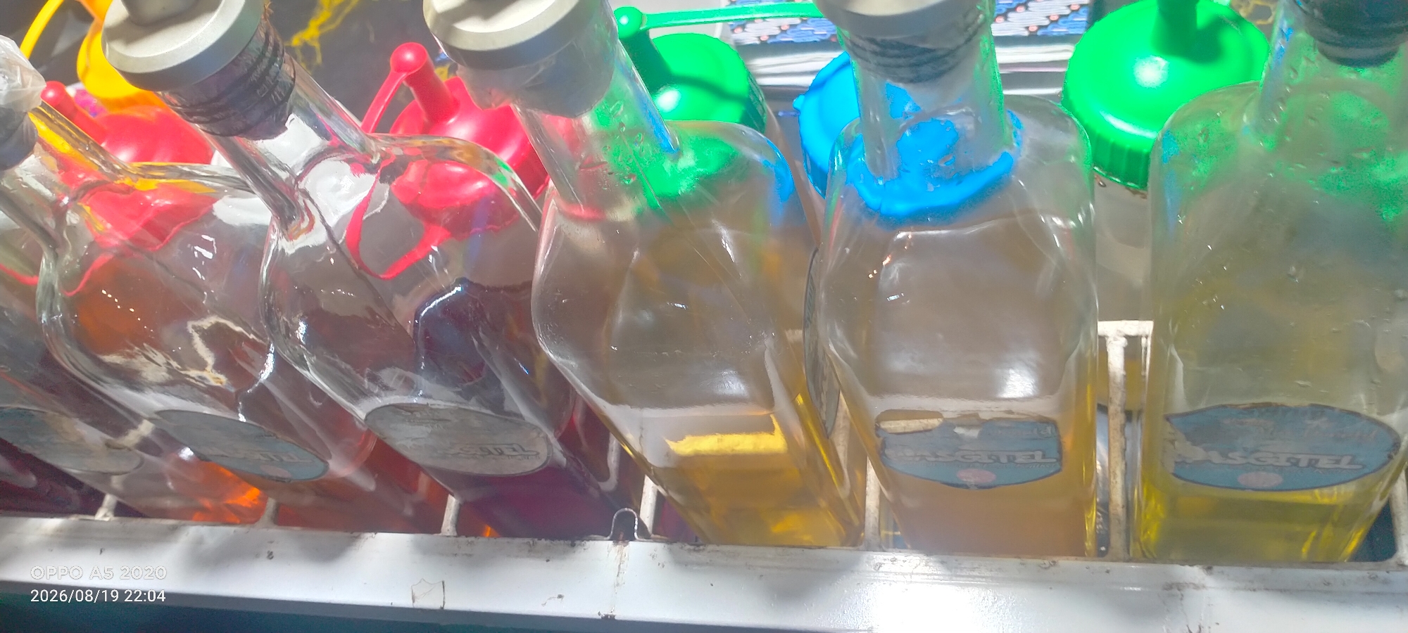 The image shows a close-up of several glass bottles with colorful caps, arranged on a shelf. The bottles appear to contain liquids of varying colors, including yellow and red. Some of the bottles have labels with the text "BASGETEL" visible. The environment appears to be a retail setting, possibly a market or store, given the arrangement of the bottles and the presence of a shelf. The bottom left corner of the image contains a timestamp and watermark indicating "OPPO A5 2020 2026/08/19 22:04". The setting is identified as Kedung, Indonesia.