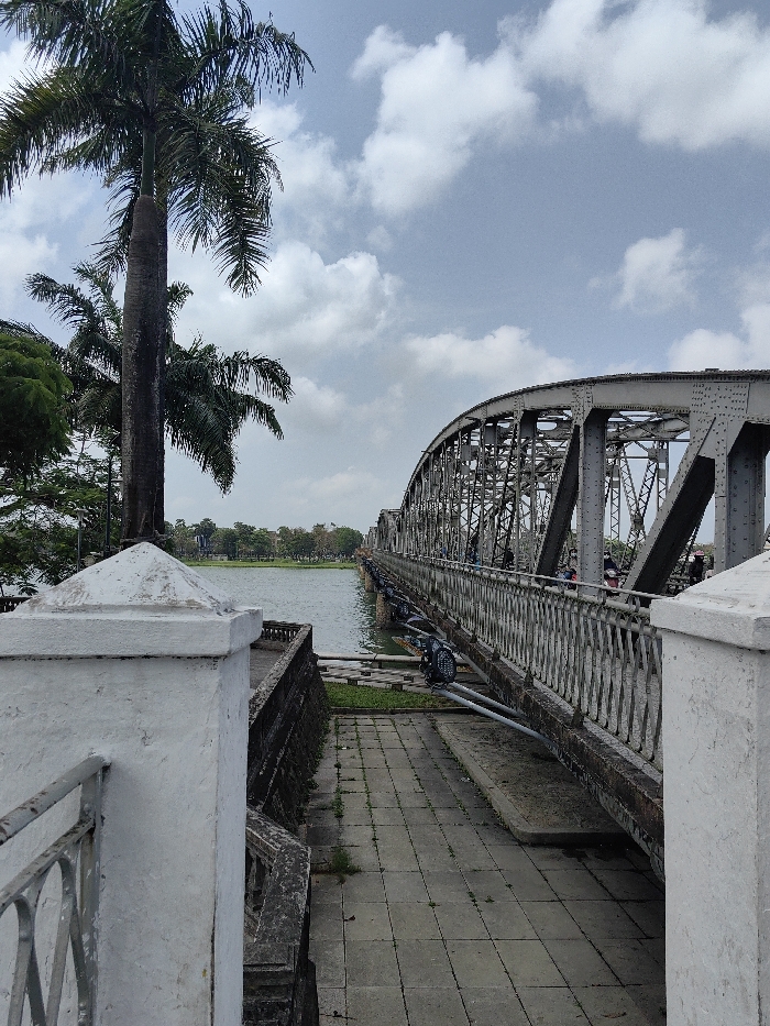The image displays an outdoor, daytime scene within Thuận Hóa Ward, Vietnam, centered on a metal truss bridge spanning a river under a partly cloudy sky. In the immediate foreground on the left, a weathered white concrete pillar with a pyramidal cap is partially visible, alongside a metal balustrade. A paved walkway composed of square concrete tiles, with some vegetative growth between the seams, extends from the foreground toward the bridge. Adjacent to the walkway on the left is a dark, rough-textured retaining wall.
To the left of the scene, a tall palm tree with a slender, segmented trunk and extensive green fronds rises prominently, with additional green foliage and other palm trees behind it. The wide river, exhibiting a dark, calm surface, occupies the central midground. Across the river, the opposite bank features green parkland or landscaped areas and distant, indistinct structures partially obscured by trees.
The primary subject is a large, grey metal truss bridge, identified as the Trường Tiền Bridge, extending from the right midground across the river towards the left background. The bridge displays an arched and riveted metal truss framework. A metal railing runs along the visible side of the bridge deck. Mounted beneath the bridge deck are several black lighting fixtures or speakers positioned on horizontal support bars. Multiple individuals are discernable on the bridge deck, some appearing to be walking, while others are on motorbikes, generally moving from the right side of the frame towards the left. The sky above is light blue with scattered white cumulus clouds.