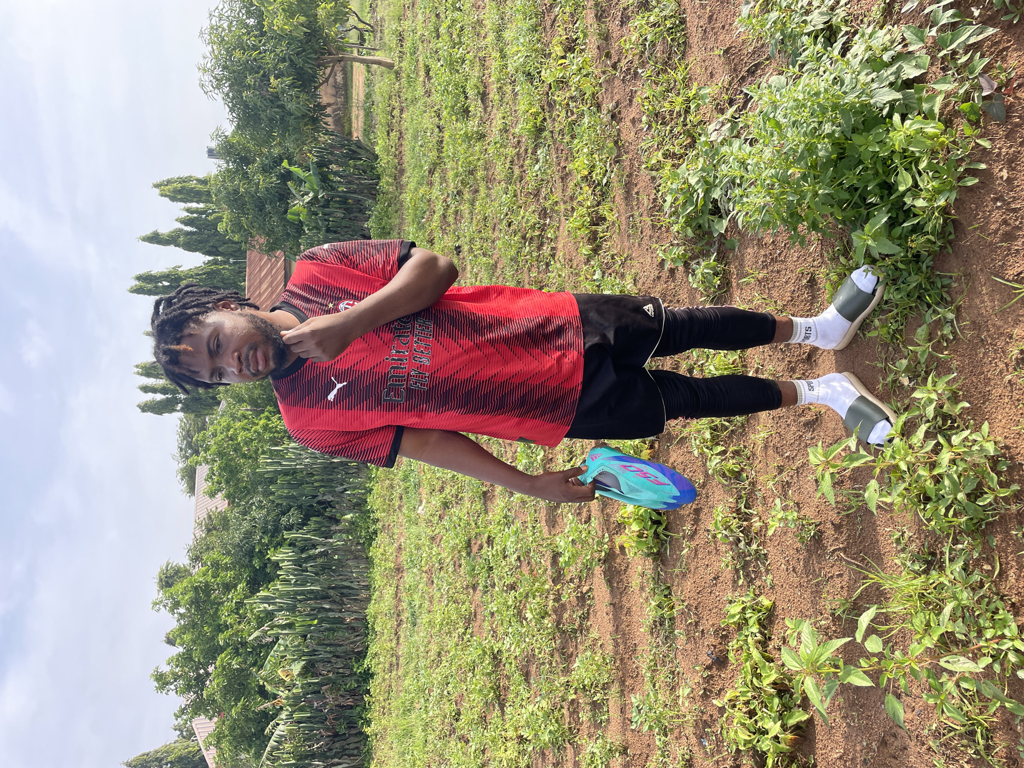 A young Black male with dreadlocks stands in a dirt field with sparse vegetation, holding a teal and blue athletic shoe in his right hand. He is wearing a red and black striped soccer jersey with "Emirates Fly Better" printed on it, black shorts over black leggings, white socks, and dark green slides. In the background, there are rows of green plants, trees, and buildings with reddish-brown roofs. The sky is overcast. The scene appears to be outdoors, possibly in a rural or suburban area, consistent with the city of Gura Top, Nigeria.