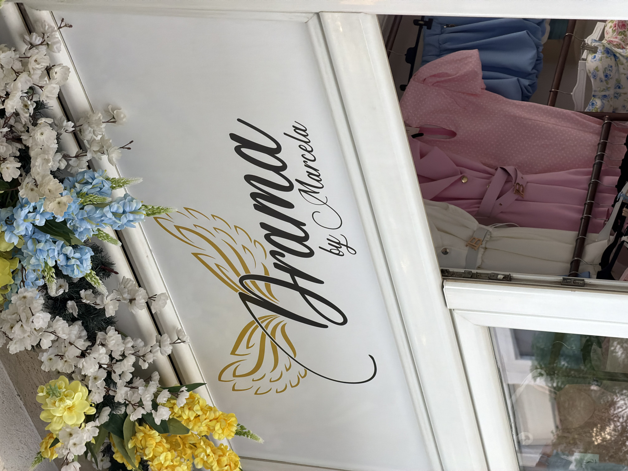 The image depicts a store window, likely in Budva, Montenegro. The window frame is white and partially obscured by floral decorations in shades of white, blue, and yellow. A sign is visible on the upper part of the window with the text "Drama by Marcela" in a decorative script. Behind the glass, clothing items hang on racks, including a pink polka-dot dress and other garments in light blue and white. The scene suggests a fashion boutique setting. The window reflects some external light, creating glare on the glass.