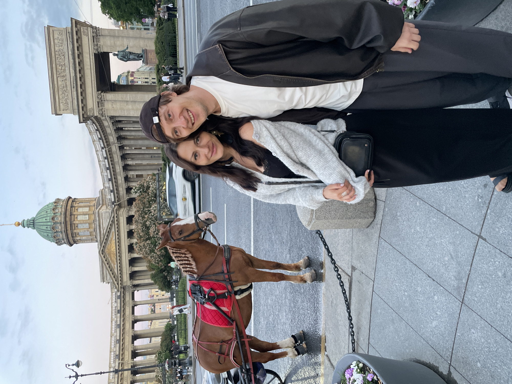 A young couple poses for a photograph in front of Kazan Cathedral in Saint Petersburg, Russia. The woman, on the left, has long dark hair and wears a black dress, a light grey cardigan, and a black crossbody bag. The man, on the right, has short brown hair and wears a black baseball cap, a white t-shirt, a black leather jacket, and grey pants. They stand close together, smiling at the camera. Behind them, a brown horse harnessed to a carriage stands on a wet paved street. The grand colonnade of Kazan Cathedral stretches across the background, topped with a green dome. Several pedestrians and a white car are visible in the distance. The scene is captured during the daytime, under a cloudy sky.