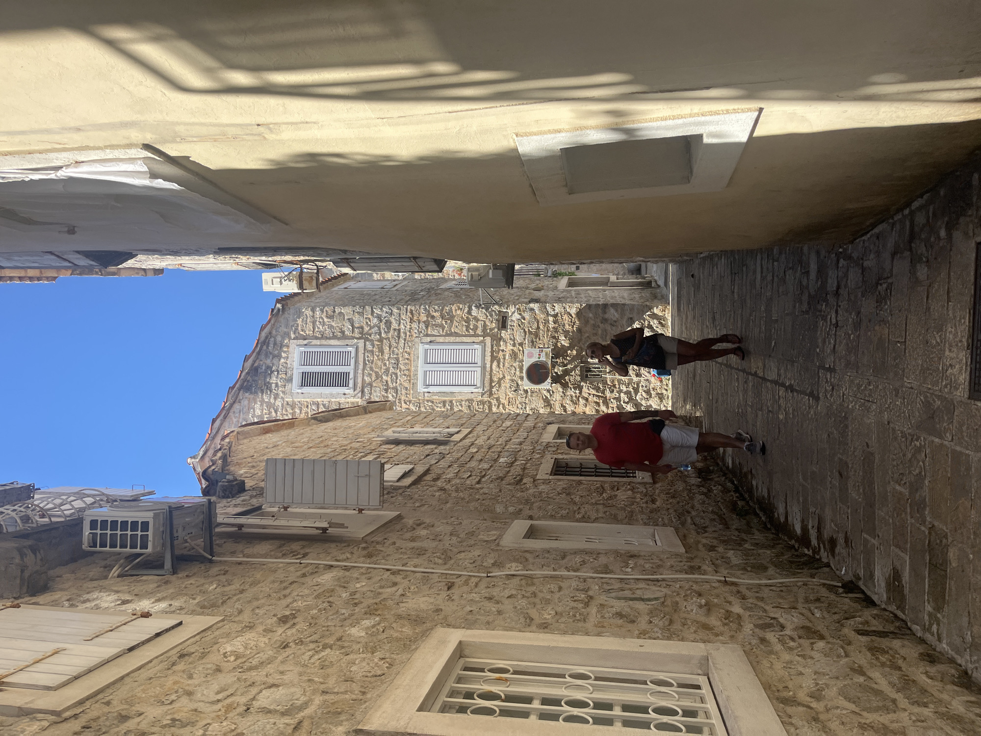 This image captures a narrow, sun-drenched alleyway in Budva, Montenegro. The scene is dominated by aged stone buildings with cream-colored facades, some featuring white-slatted shutters. The architecture suggests a historic European coastal town. The sky is a vibrant, clear blue, indicating fair weather and likely midday.
Two individuals are visible in the alley. The first, wearing a dark tank top and shorts, is walking away from the camera towards the right side of the frame. The second, a person in a red t-shirt and white shorts, is standing and looking towards the left. Their presence suggests a leisurely stroll or exploration of the area.
Notable details include the presence of air conditioning units mounted on the exterior walls of the buildings, indicating modern amenities within the historic setting. Shadows cast by overhead structures create a dynamic play of light and shade across the stone pavement and walls. The overall mood is relaxed and summery, typical of a tourist destination. No visible text is present in the image.