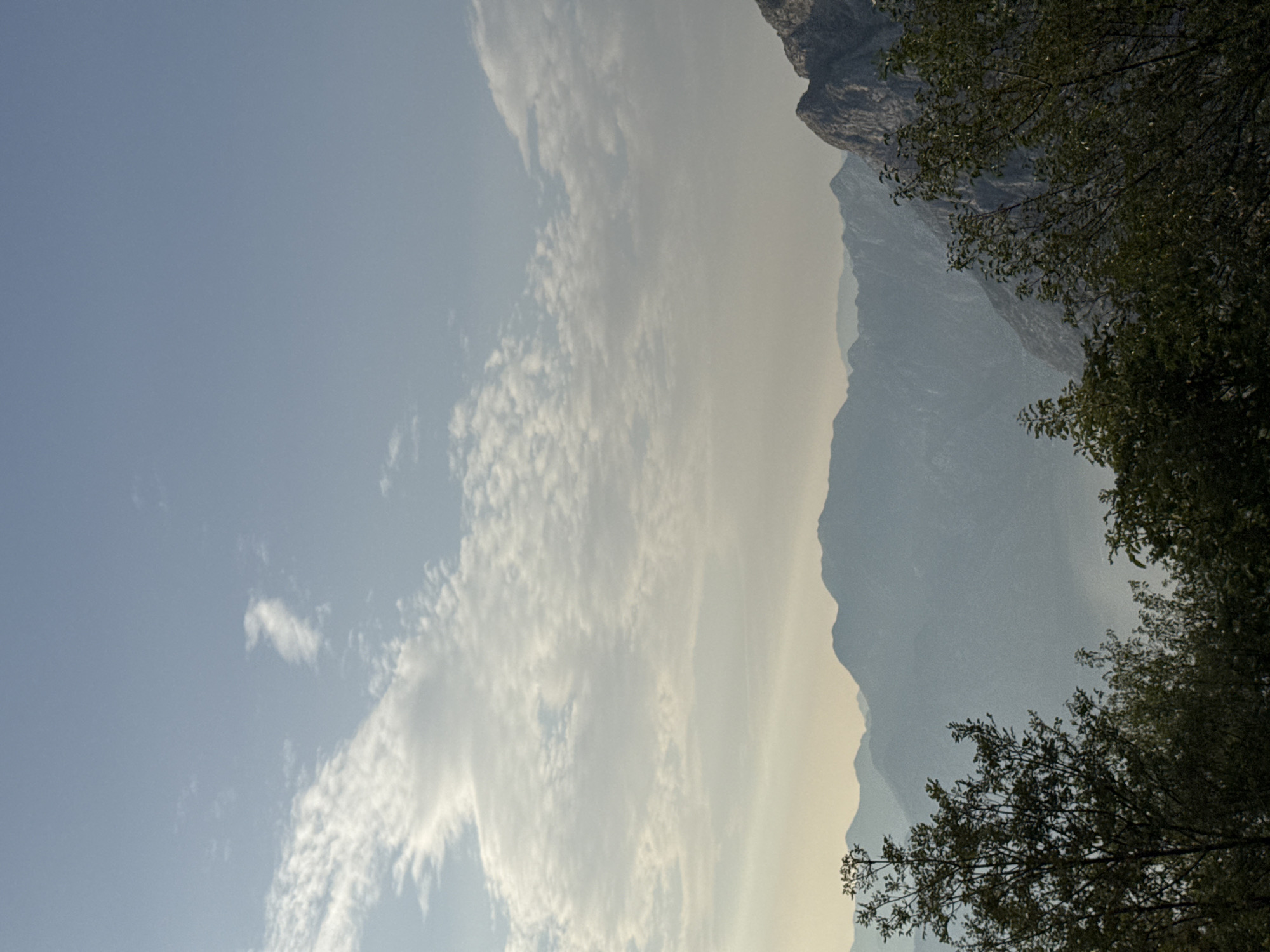 This media file captures a natural landscape with a focus on mountains and sky. The scene is dominated by the majestic, hazy outlines of mountains in the background, which recede into the distance, creating a sense of depth. In the foreground, the dark, leafy branches of trees frame the right side of the image, providing a contrast to the lighter sky.
The sky is a soft, pale blue, partially obscured by wispy, elongated clouds that stretch across a significant portion of the frame. The light suggests it is either early morning or late afternoon, with a gentle, diffused illumination. The haze in the mountains indicates a warm day or potentially atmospheric conditions common in mountainous regions.
There are no people or discernible objects other than the natural elements of trees, mountains, and clouds. The overall mood is serene and peaceful, emphasizing the vastness and quiet beauty of the natural world. The location context of Žanjev Do, Montenegro, points to a mountainous, possibly rural or wilderness area. No text is visible in the image.