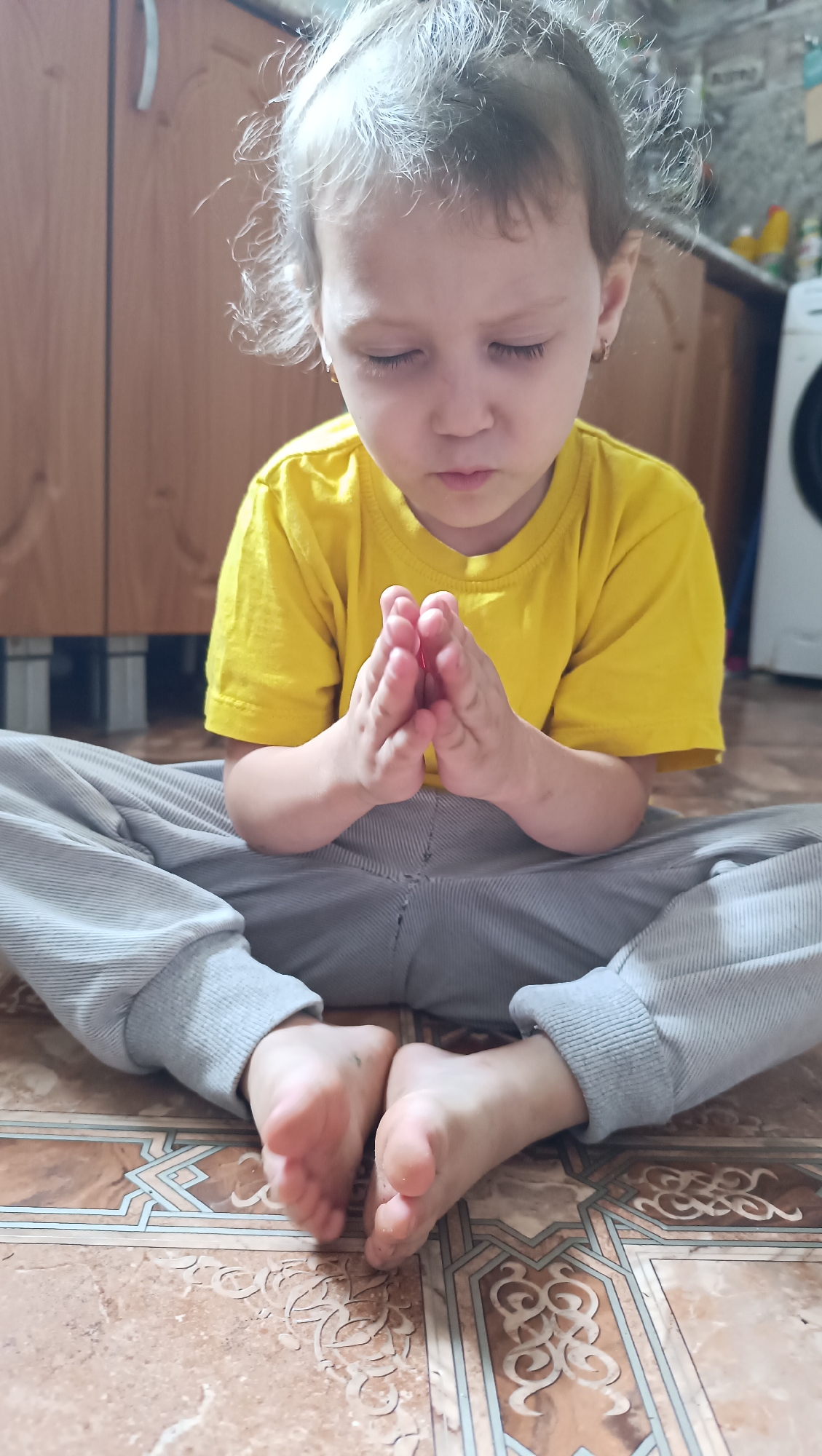 A young Caucasian child with blonde hair is sitting on the floor with their legs crossed and hands pressed together in front of their chest. The child is wearing a yellow t-shirt and grey sweatpants. The child's eyes are closed. The floor is tiled with a patterned design. In the background, there are wooden cabinets and a white appliance, possibly a washing machine. The scene appears to be indoors, likely in a home. The location is identified as the city of Karsha, Russia.