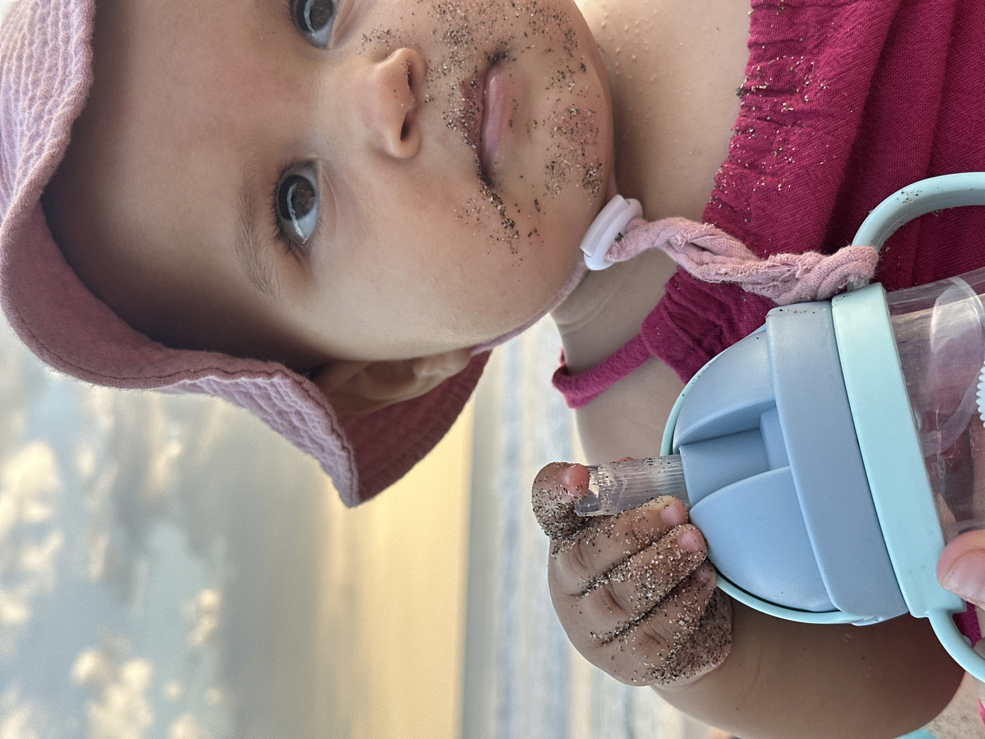 A low-angle, medium shot captures a baby, face covered in sand, wearing a pink hat and shirt, and holding a light blue and clear bottle. The background is blurred with sand visible. The location is North Kuta, Indonesia, as evidenced by the beach setting. The subject appears to be playing on a beach. The baby is holding the bottle in their sandy hand.