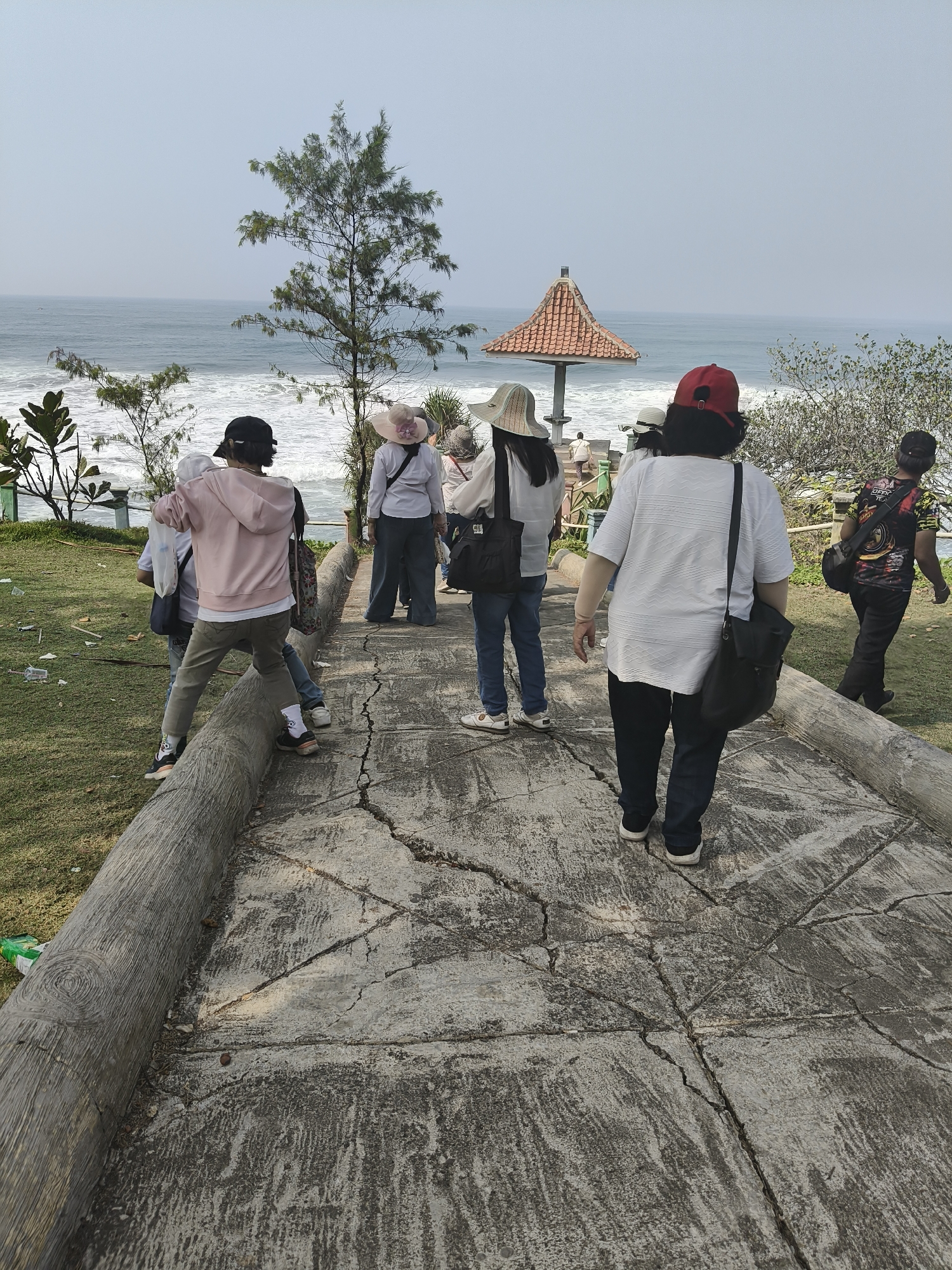 In Desa Ciliang, Indonesia, a group of people are walking on a concrete path beside a grassy area. The path is lined with a log-like barrier. In the background, the ocean is visible with waves crashing on the shore. A small pavilion with a tiled roof is situated near the path. The people are dressed in casual clothing, with some wearing hats. The atmosphere appears to be relaxed and leisurely, suggesting a scenic outing or a visit to a coastal attraction.