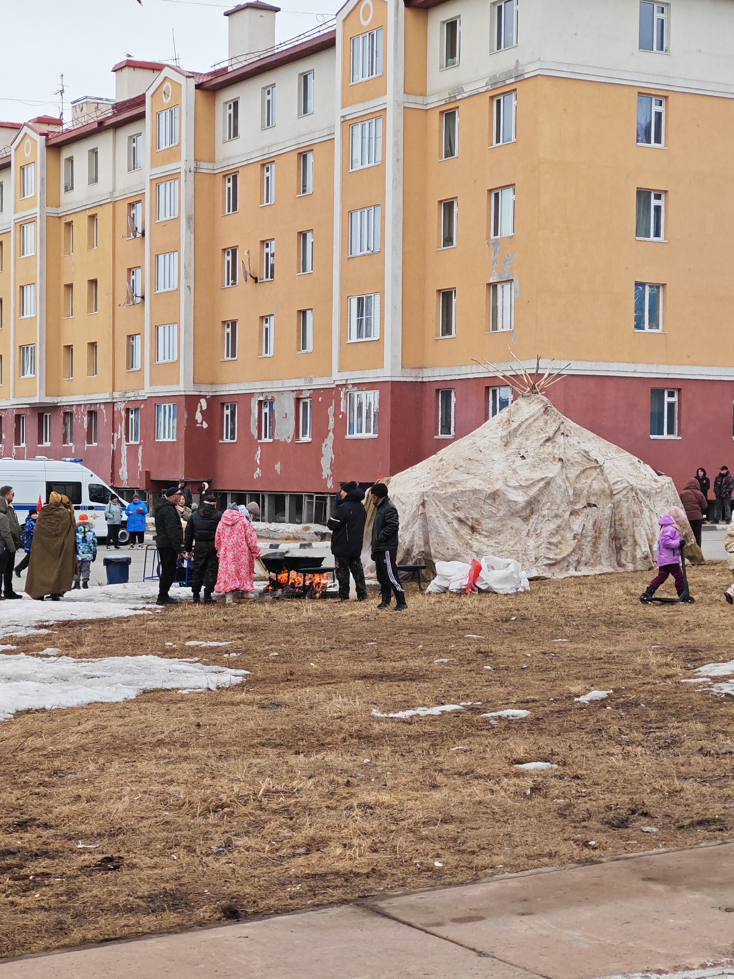 In Ugolnye Kopi, Russia, a group of people stand around a large open fire set in a blackened metal container on a field of dead grass with patches of melting snow. The group consists of roughly 10-15 individuals, some in warm coats. A large, beige, teepee-like structure is positioned behind them. To the left, an ambulance is visible, with additional individuals nearby. A multi-story apartment building with orange and white facades forms the backdrop. A child in a purple coat rides a scooter to the right of the teepee. Two white sacks are adjacent to the teepee.