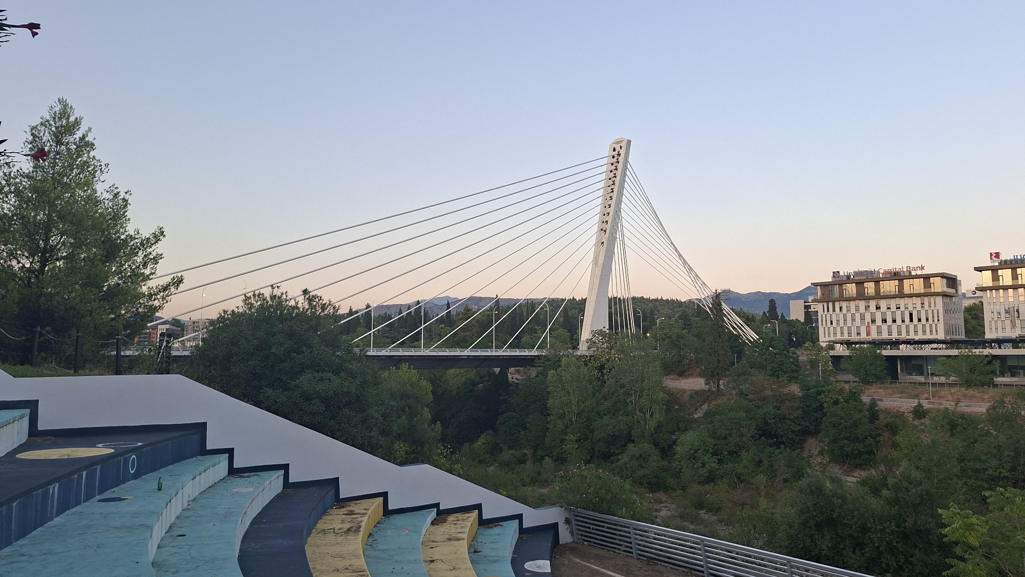 This is a daytime, eye-level, wide-angle shot of an outdoor amphitheater in Podgorica, Montenegro, with a modern cable-stayed bridge and city buildings in the background. The foreground features a section of an amphitheater with curved rows of seats painted in blue, yellow, and dark gray. The seats appear worn. Behind the amphitheater is a dense area of green trees and foliage, leading to a riverbed or ravine.
In the mid-ground, a prominent white cable-stayed bridge spans the ravine. The bridge has a single, tall, triangular pylon from which numerous cables extend to support the bridge deck. Further in the background, across the bridge, are several modern, multi-story buildings. The building on the right has signage that reads "Capital Bank" and "Hotel". The sky is clear and light blue, transitioning to a warmer hue near the horizon, suggesting either early morning or late afternoon. There are no people visible in the image.