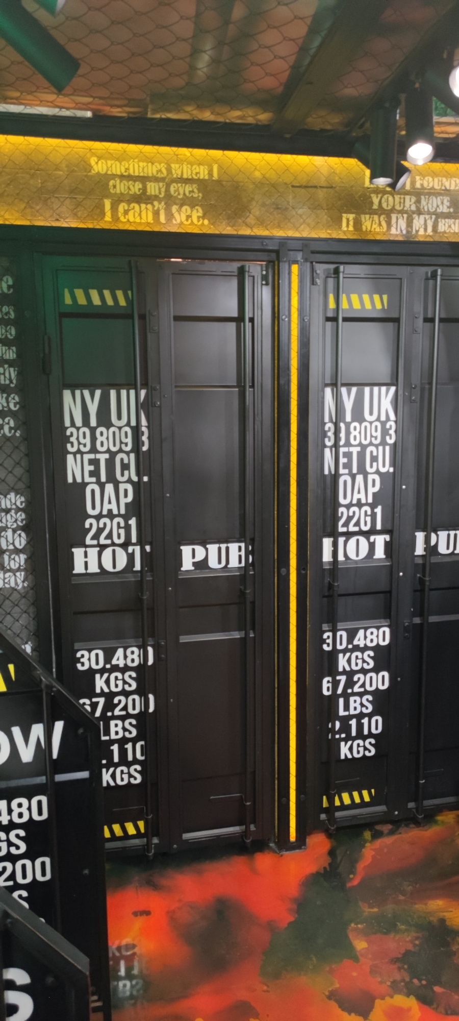 The image depicts the interior of a commercial establishment, likely a restaurant or bar, with a industrial-themed decor. Prominently featured are large, dark-colored doors resembling shipping containers, some of which have stenciled text and numbers in white. The text includes "NY UK", "NET CU.", "OAP", "22G1", "30.480 KGS", "67.200 LBS", and "2.110 KGS". One door also has the partial word "PUB" visible. Above the doors, a yellow illuminated sign displays text in white lettering. The left side of the sign reads "Sometimes when I close my eyes, I can't see." The right side of the sign is partially obscured but appears to contain text related to being found, a nose, and possibly a business.
The floor is covered in a decorative pattern resembling spilled paint or abstract art, with vibrant red, orange, and dark green colors. The ceiling is a dark mesh material with industrial lighting fixtures. The overall ambiance suggests a modern, edgy aesthetic. There are no people visible in the image. The establishment is located in the city of Yerevan, Armenia.