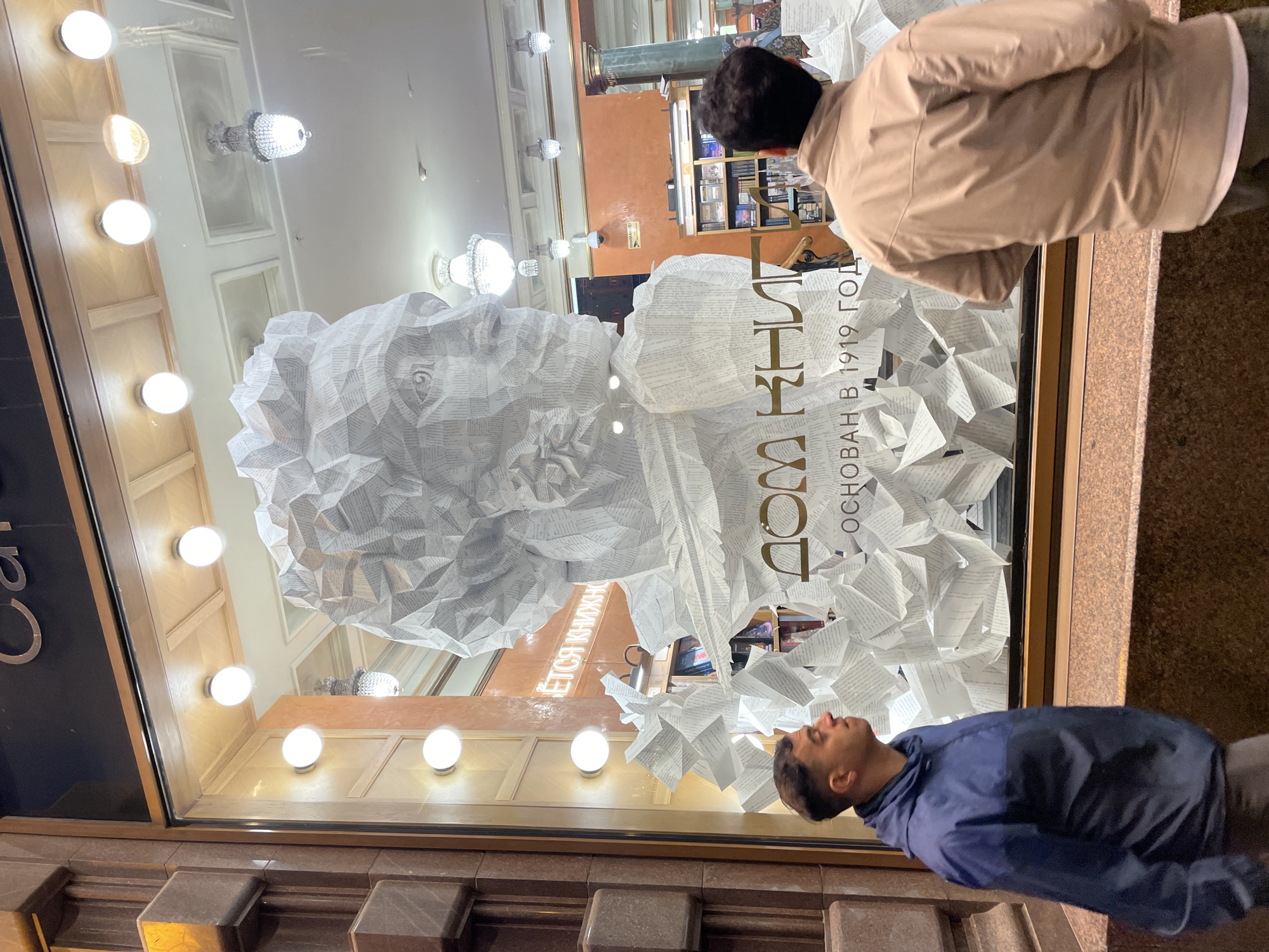 Two men are captured at night, looking into the illuminated window display of a bookstore. The image is oriented vertically, showing the street level at the bottom and the top of the shop window at the top. The man in the foreground, wearing a dark blue jacket, is looking up and into the display. Behind him, another man in a beige jacket also observes the window.
The shop window features a striking, large-scale bust or sculpture made entirely from countless folded white paper pages, creating a three-dimensional, textured surface. This paper artwork dominates the display, with additional folded paper elements scattered around its base, giving the impression of an abstract literary landscape.
Prominently displayed on the window in gold Cyrillic lettering is "ДОМ КНИГИ" (Dom Knigi), which translates to "House of Books" or "Bookstore." Below this, "ОСНОВАН В 1919 ГОД" (Osnovan v 1919 god) is visible, indicating the store was "Founded in 1919." Partial text to the left reads "ЕТСЯ КНИЖКУ," likely part of a longer phrase related to books.
Through the window, the interior of the bookstore is visible, showcasing warm lighting, bookshelves filled with books, and decorative chandeliers. The exterior facade of the building appears to be made of dark brown stone or tile. The overall impression is one of an engaging and artistic window display attracting passersby in the evening.