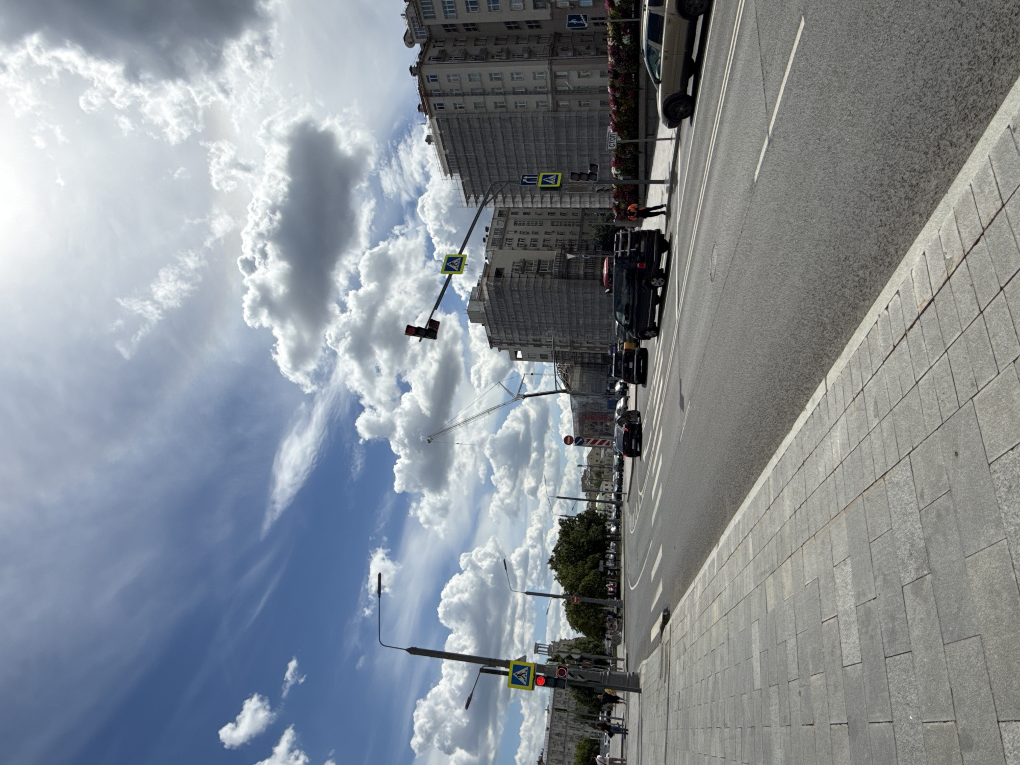 The image shows a street scene in a city with a blue sky filled with white and gray clouds. A paved road with lane markings runs diagonally from the bottom right to the top left. Several cars are driving on the road, including a black SUV and a light-colored SUV. A pedestrian walkway with gray paving stones is visible on the left side of the road. Traffic lights and streetlights are present. Buildings with scaffolding are seen in the background, indicating construction. There are also trees and some flowers. A few people are walking on the pedestrian walkway. The overall impression is of a busy urban environment on a partly cloudy day.