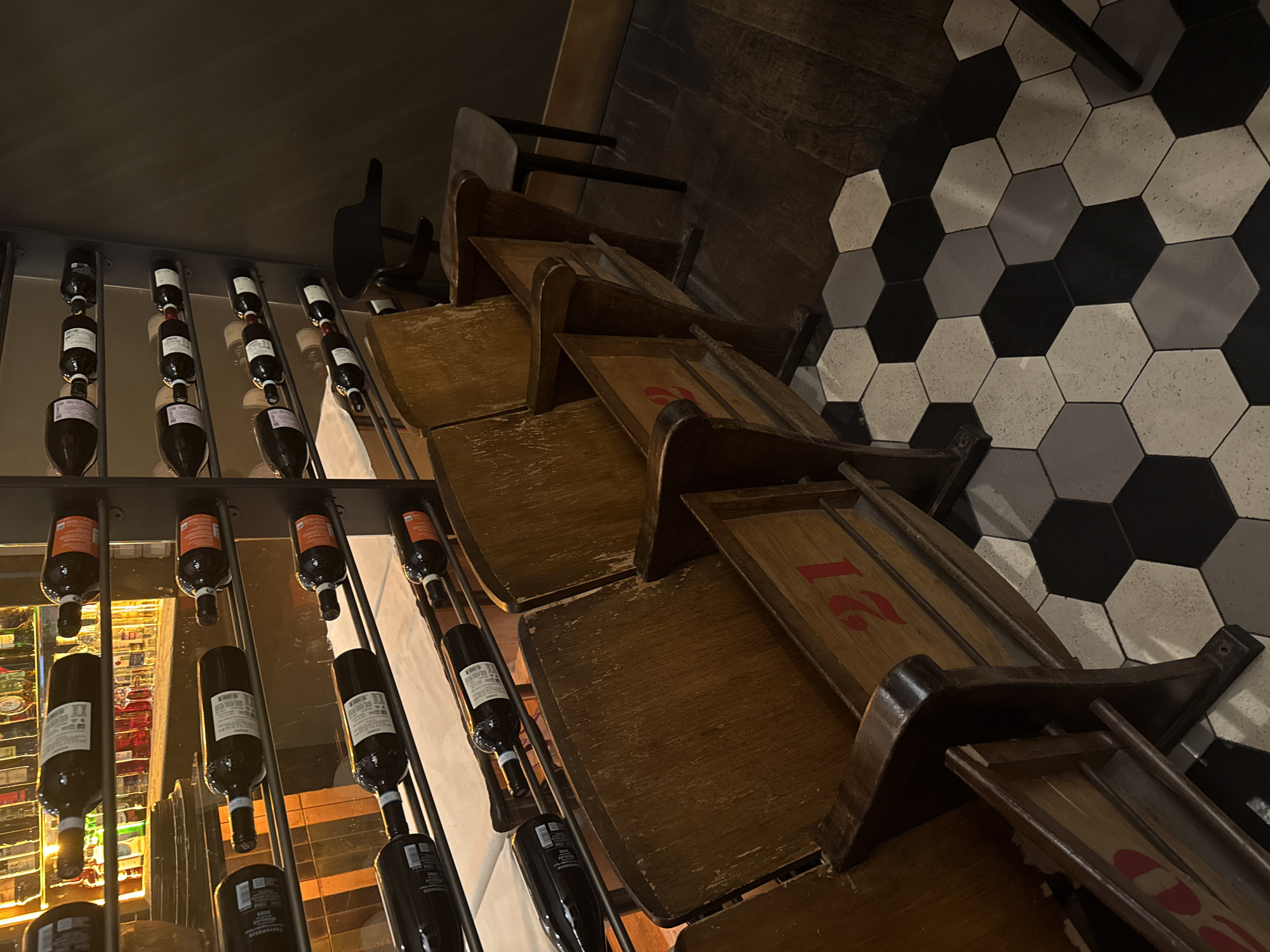 The image depicts a detail of a bar in Milan, Italy. A rack displaying wine bottles sits alongside a row of antique, wooden chairs. The floor is tiled with black and white hexagons. The lighting is dim and warm.