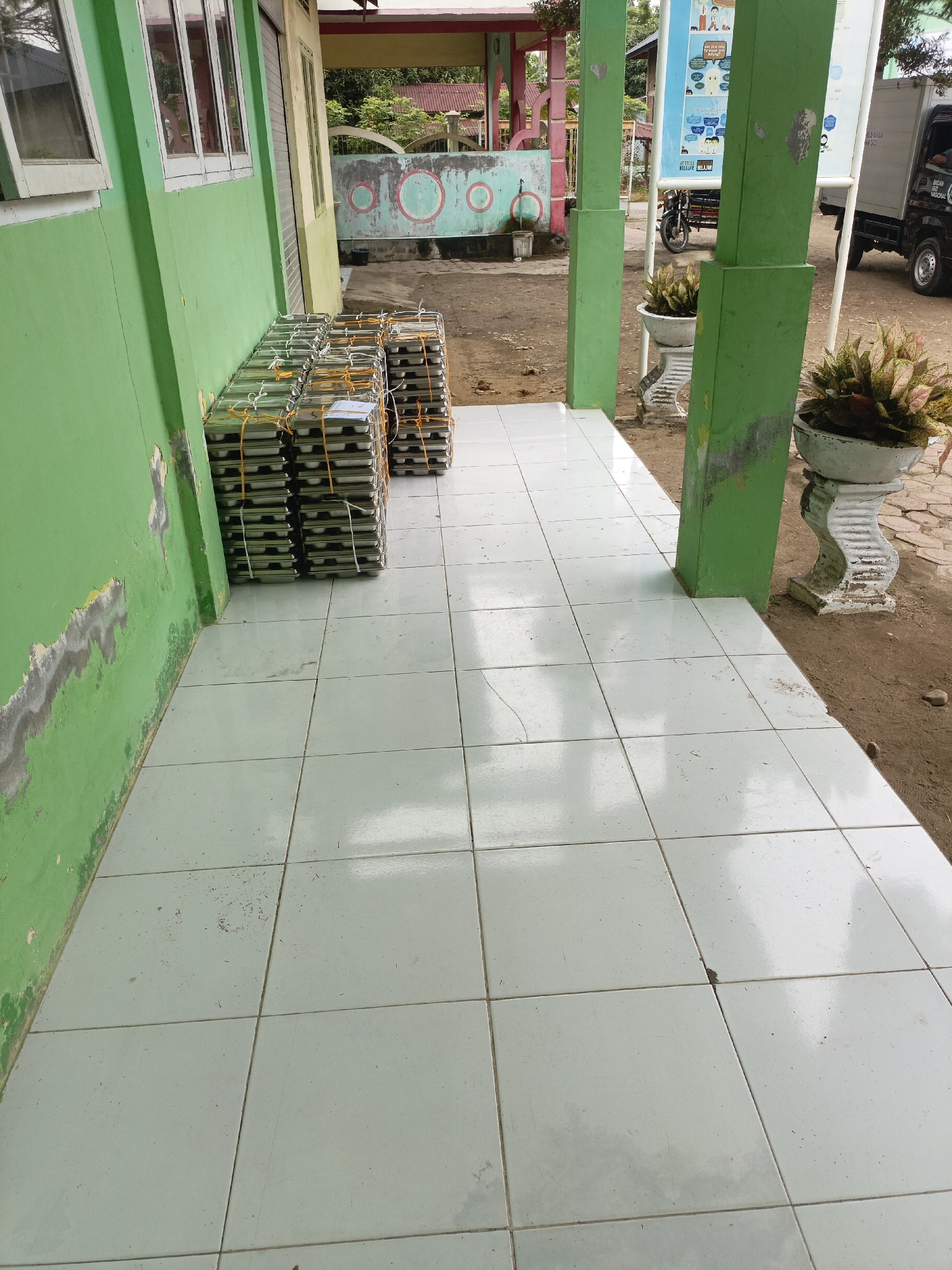 The image shows stacks of styrofoam containers, likely food trays, lined up along the exterior wall of a building. The building's wall is painted green and shows signs of wear and tear. The containers are wrapped in plastic and secured with orange string. They are arranged in neat rows, suggesting preparation for an event or distribution.
In the background, there is a white banner with text and illustrations, a motorcycle, and a white truck. Two potted plants are visible, one on a pedestal. The ground is a mix of dirt and paved tiles. The overall scene appears to be outdoors, possibly in a school or community center setting, given the banner and the outdoor area. The lighting suggests it is daytime. The location context of Lhokseumawe, Indonesia, indicates a tropical climate. There is no discernible activity or event taking place other than the arrangement of the food containers. The visible text on the banner is too blurry to be read.