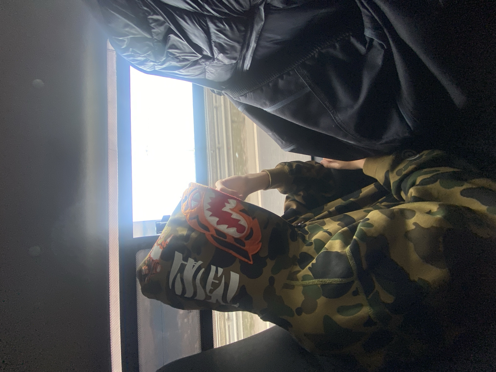A person wearing a camouflage hoodie with a shark design on the hood sits beside a window. A dark garment hangs partially over the window frame. The location is not discernible from the image.