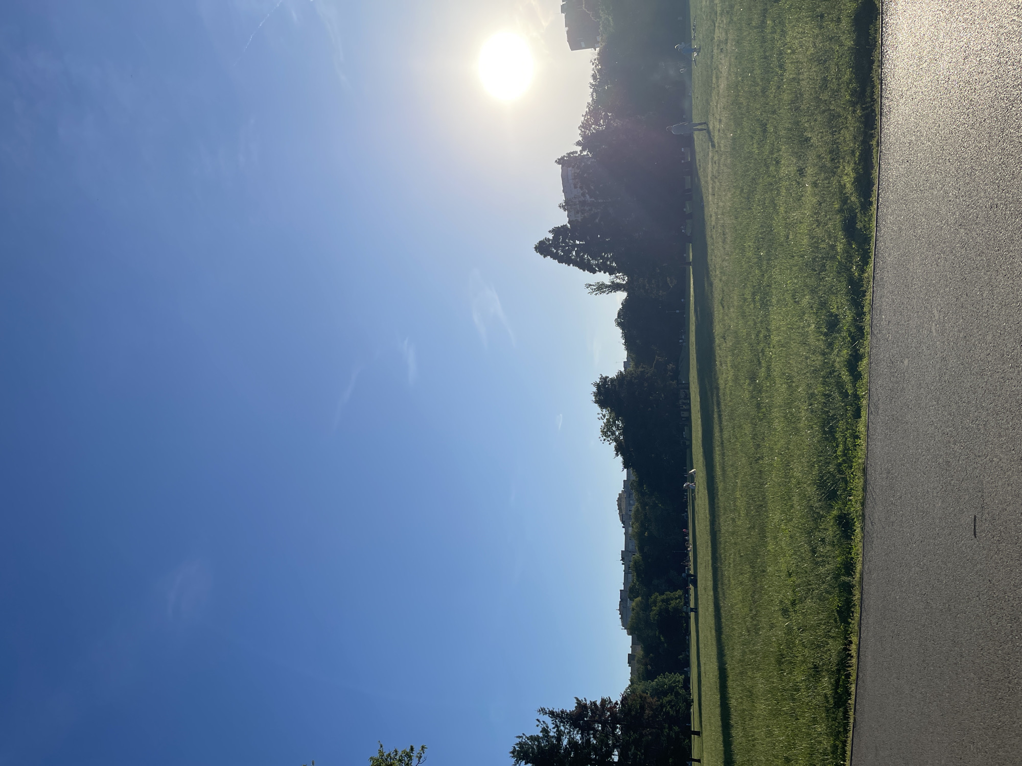 The image captures a sunny day in a park, likely located in Sofia, Bulgaria due to the presence of architecture and vegetation common to the region. The sky is predominantly clear blue with a few wispy clouds. The sun is high, positioned towards the upper center of the frame. Below the sky is a grassy park. A paved pathway runs along the side of the grass. In the distance, there is a treeline and buildings. There are several people enjoying the park, some are walking, some are standing.