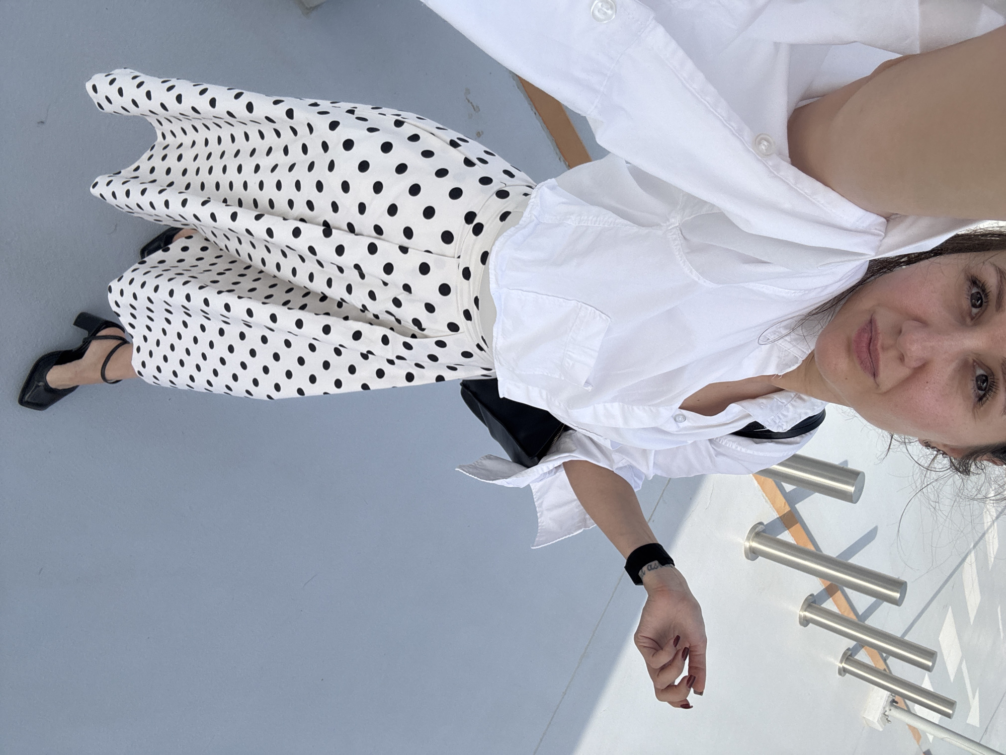 A full-body selfie captures a woman standing outdoors on a light grey-blue paved surface, looking directly at the camera with a calm expression. The image is rotated, presenting her at an angle.
She is dressed in a classic black and white outfit. She wears a crisp white long-sleeved button-down shirt, partially unbuttoned at the neckline. Below, she sports a flowing white skirt with an all-over pattern of various-sized black polka dots. On her feet, she wears black block-heeled shoes with straps. A small black handbag or clutch is partially visible tucked under her arm. Her left wrist features a black wristband, under which a tattoo is visible, displaying the partially discernible text "I am as". Her fingernails are painted a dark red.
The background shows more of the paved ground, with strong shadows cast by unseen objects, indicating bright, sunny daytime conditions. In the distance, several silver-colored, cylindrical metallic posts or bollards are arranged in a line, also casting long shadows. The scene suggests an outdoor public area, possibly in Dubai, United Arab Emirates, given the provided context.