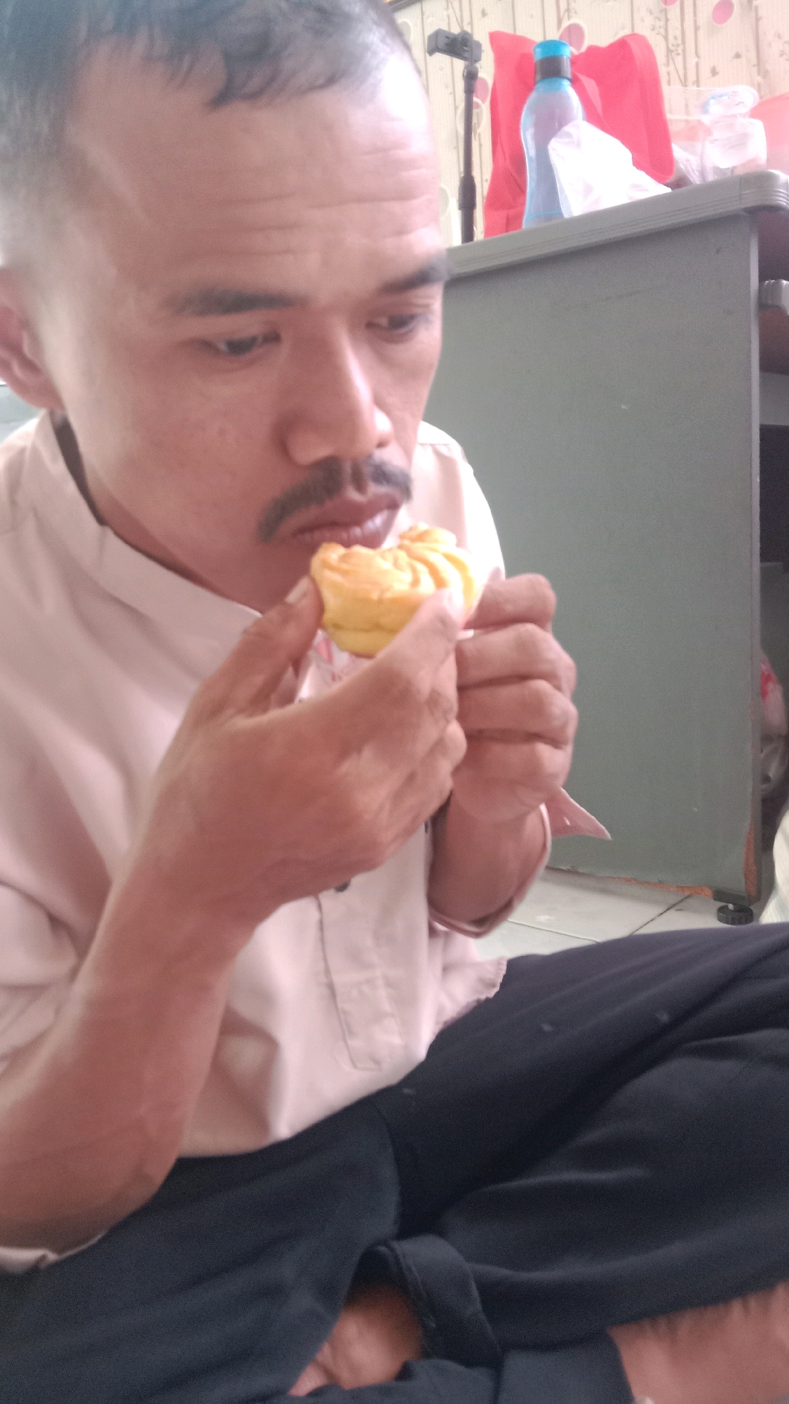 A man in a light pink collared shirt and dark pants is sitting down, holding a small, yellow, pastry-like food item up to his mouth. He has short, dark hair, a mustache, and is looking down at the food. His right hand is holding the pastry, and his left hand is near it. In the background, on a green surface, is a blue water bottle and a red object. The scene appears to be indoors.