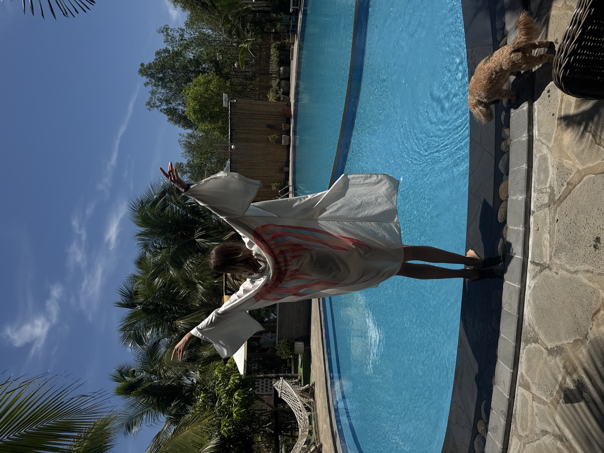 In Mũi Né, Vietnam, a young woman stands with her back to the camera, facing a bright blue swimming pool. She is wearing a flowing white kimono-style garment adorned with a large, stylized pink and red wing design on the back. Her arms are outstretched, palms facing outwards, as if embracing the sunny day. She is barefoot and her legs are visible below the garment. Beside the pool, a small, fluffy light brown dog is looking towards the water. The pool area is surrounded by stone paving and lush green palm trees. In the background, a hammock hangs from wooden posts, and a bamboo fence lines the property. The sky is a clear, vibrant blue with a few wispy clouds.