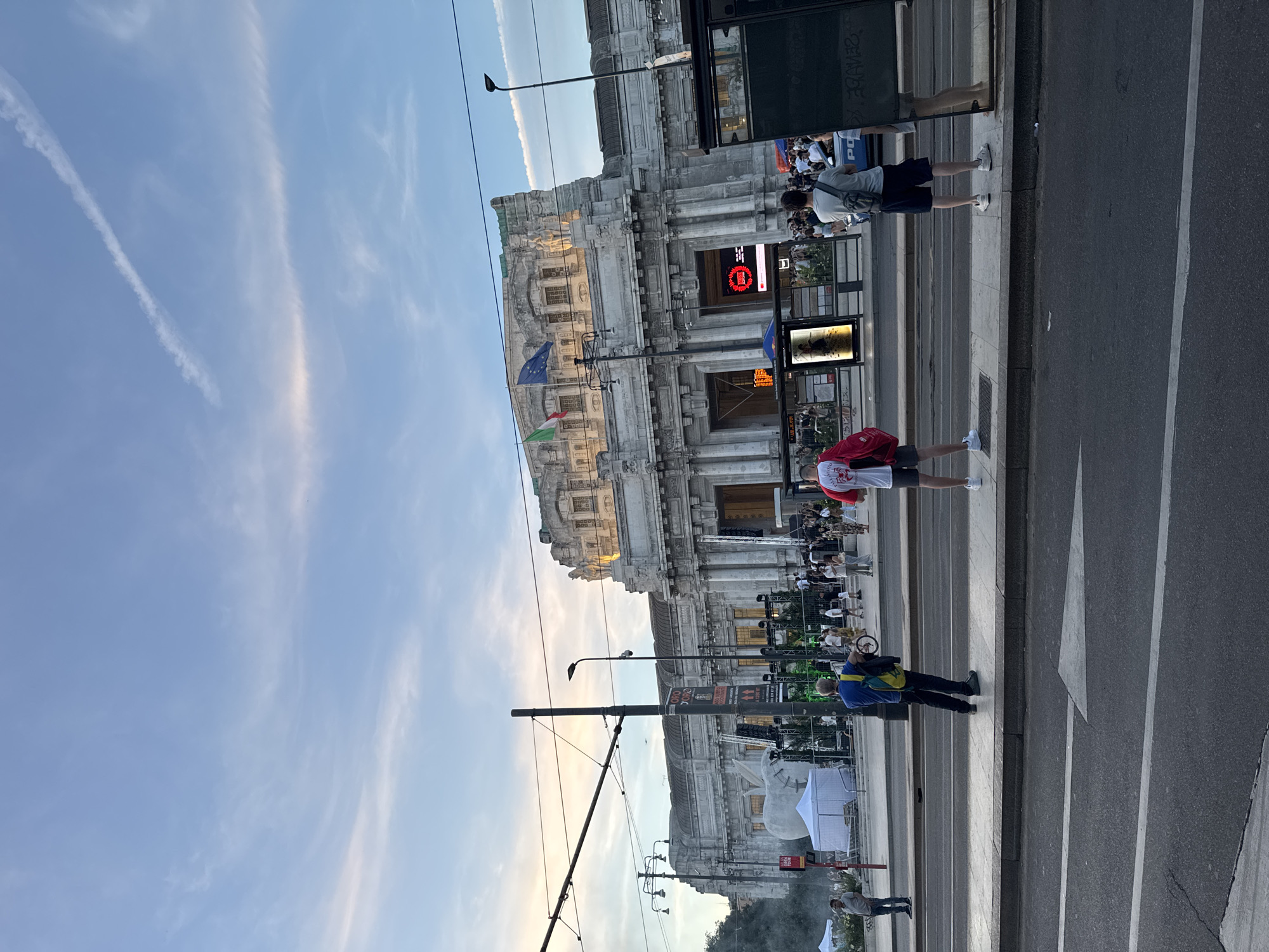 This is a medium shot, eye-level, full-frame photograph of the Milan Centrale train station in Milan, Italy, taken from across the street. The sky is a pale blue with wispy clouds, some of which are tinged with pink from the setting sun. A contrail stretches across the sky from upper left to lower right.
The grand, neoclassical facade of the train station dominates the background. It is a large, imposing stone building with multiple stories, adorned with columns, statues, and architectural details. Two flags, one Italian and one European Union flag, fly from poles on the upper part of the building. In front of the station, there are stages, tents, and various temporary structures set up, suggesting an event or exhibition is taking place. A large, white, abstract sculpture is visible in the middle ground.
In the foreground, there is a wide street with tram tracks. A sidewalk runs along the street, and several people are standing on it, waiting or observing. On the sidewalk, there are bus stop shelters with advertisements.
To the left, a man in a gray shirt and blue jeans stands near a white tent. In the center, a man with a blue shirt and black pants, carrying a yellow and green bag over his shoulder, stands with his back to the camera. To the right, a man wearing a white t-shirt with red lettering and dark shorts stands with his back to the camera, holding a red bag. Further right, another man in a gray t-shirt and dark shorts is also visible. Other people are scattered across the sidewalk and in front of the station, mostly appearing to be either spectators or participants in the event.
The lighting suggests it is late afternoon or early evening, with the sun beginning to set. The overall scene depicts a public space in Milan bustling with activity around a significant architectural landmark.