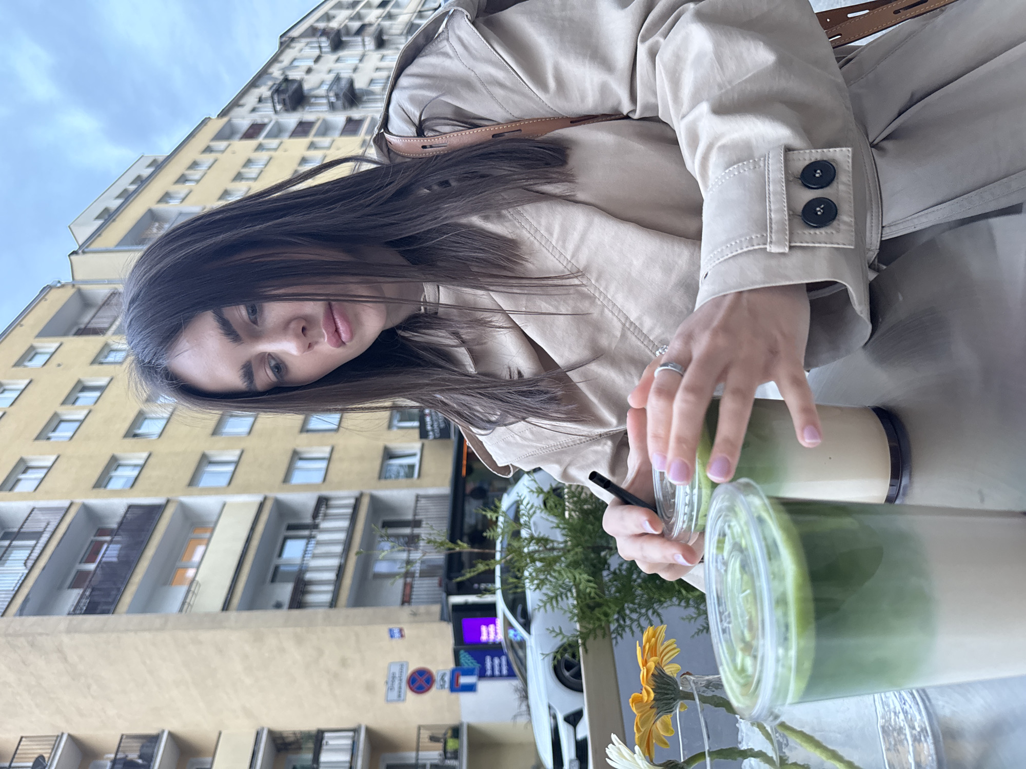 A woman with brown hair is seated outdoors, likely at a cafe or restaurant, with two drinks in front of her. The drinks appear to be matcha lattes, layered green and tan, in clear plastic cups with lids. One drink has a black straw inserted. The woman is wearing a tan trench coat.
Behind her is a multi-story apartment building, yellow in color. Street signs are visible. A white car and greenery add depth to the scene. This photograph was taken in the city of Warsaw, Poland.