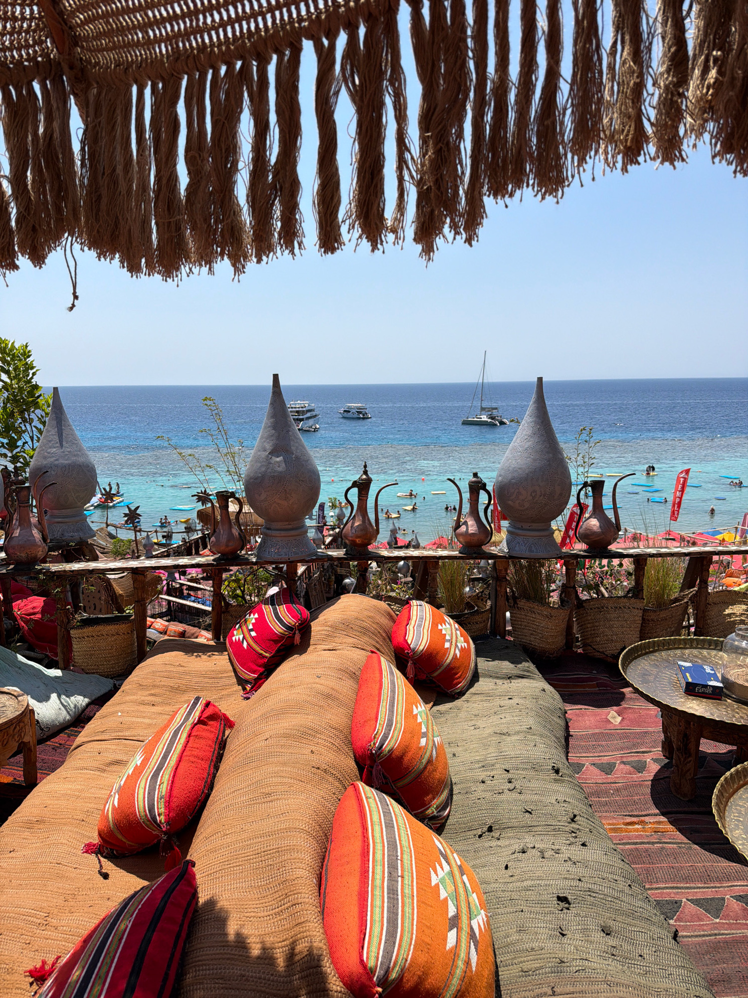 This image depicts an outdoor seating area at a beach resort in Sharm El Sheikh, Egypt, overlooking the Red Sea. The foreground is dominated by a comfortable seating arrangement with large, woven floor cushions and colorful throw pillows adorned with geometric patterns in red, orange, black, and white. Behind the seating area, a wooden railing separates the space from the beach and the sea. Various decorative items, including large, metallic urns and copper pitchers, are placed along the railing.
In the middle ground, a vibrant beach scene unfolds. People are visible on the sand, in the shallow turquoise water, and on various watercraft, including paddleboards and small boats. Several larger boats are anchored further out in the blue sea. Red flags are planted along the beach. The water is crystal clear, revealing coral formations beneath the surface.
The background consists of a clear, bright blue sky. Above the seating area, a thatched canopy with hanging rope fringe provides shade. The overall atmosphere is one of a relaxed, tropical vacation destination. The resort appears to be situated in a coastal area known for its clear waters and coral reefs, characteristic of Sharm El Sheikh's popular dive and snorkeling sites.