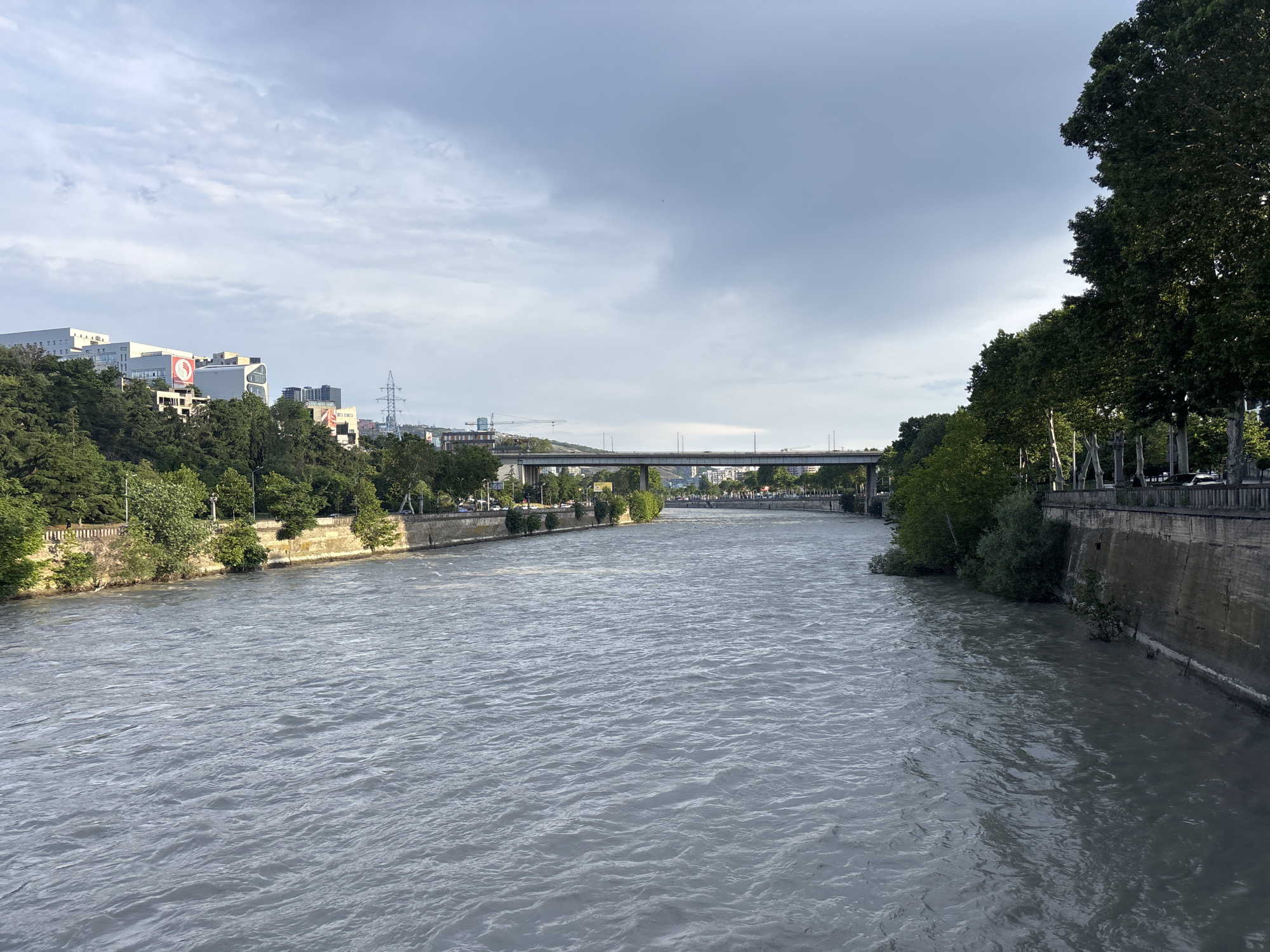 The image depicts a wide, murky river flowing through the city of Tbilisi, Georgia. On the left bank, there are densely vegetated hills dotted with modern buildings and a large red and white circular logo. Further along the left bank, a long, multi-lane bridge spans the river, with traffic visible on it. The right bank is characterized by a stone embankment lined with a row of mature trees casting shadows. The sky above is overcast with scattered clouds. The overall scene is a cityscape with a prominent river as the central element, likely captured from another bridge or elevated viewpoint.