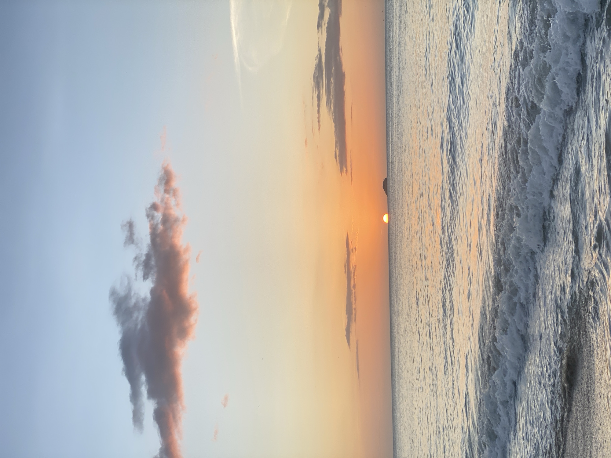 This is a photograph of a sunset over the ocean in Pedro González, Venezuela. The sun is a small, glowing orb just above the horizon, casting a warm, orange hue across the sky. A few wispy clouds are scattered across the sky, tinged with pink and orange. The ocean is calm, with gentle waves lapping at the shore. In the distance, a small island is visible. The overall mood of the image is peaceful and serene.