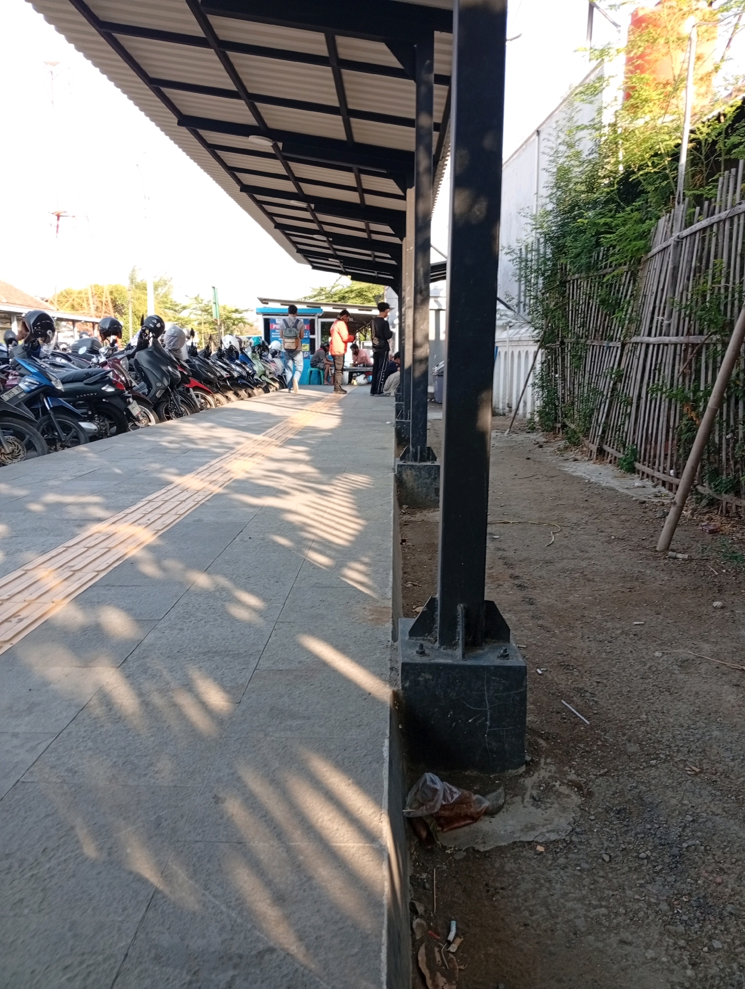 This image depicts an outdoor scene at what appears to be a transportation hub or waiting area, possibly a bus or train station, in Karawang, Indonesia. The foreground features a paved walkway with a textured tactile paving strip running along its length, likely to assist visually impaired individuals. A row of parked motorcycles is visible to the left, partially shaded by a corrugated metal roof supported by dark metal pillars. Sunlight casts long shadows across the walkway from the roof structure.
In the mid-ground, several people are present. Some are standing near what looks like a ticket booth or information kiosk, with one individual in an orange shirt and another in jeans carrying a backpack. A few other individuals are seated on a bench or low wall. Beyond the parked motorcycles, there are hints of buildings and vegetation.
The right side of the image shows a dirt and gravel area next to a fence constructed of vertical wooden poles. Green foliage grows along the fence line. A few pieces of trash are visible on the ground near the base of a metal pillar in the right foreground. The overall impression is one of a functional, everyday public space with activity.
