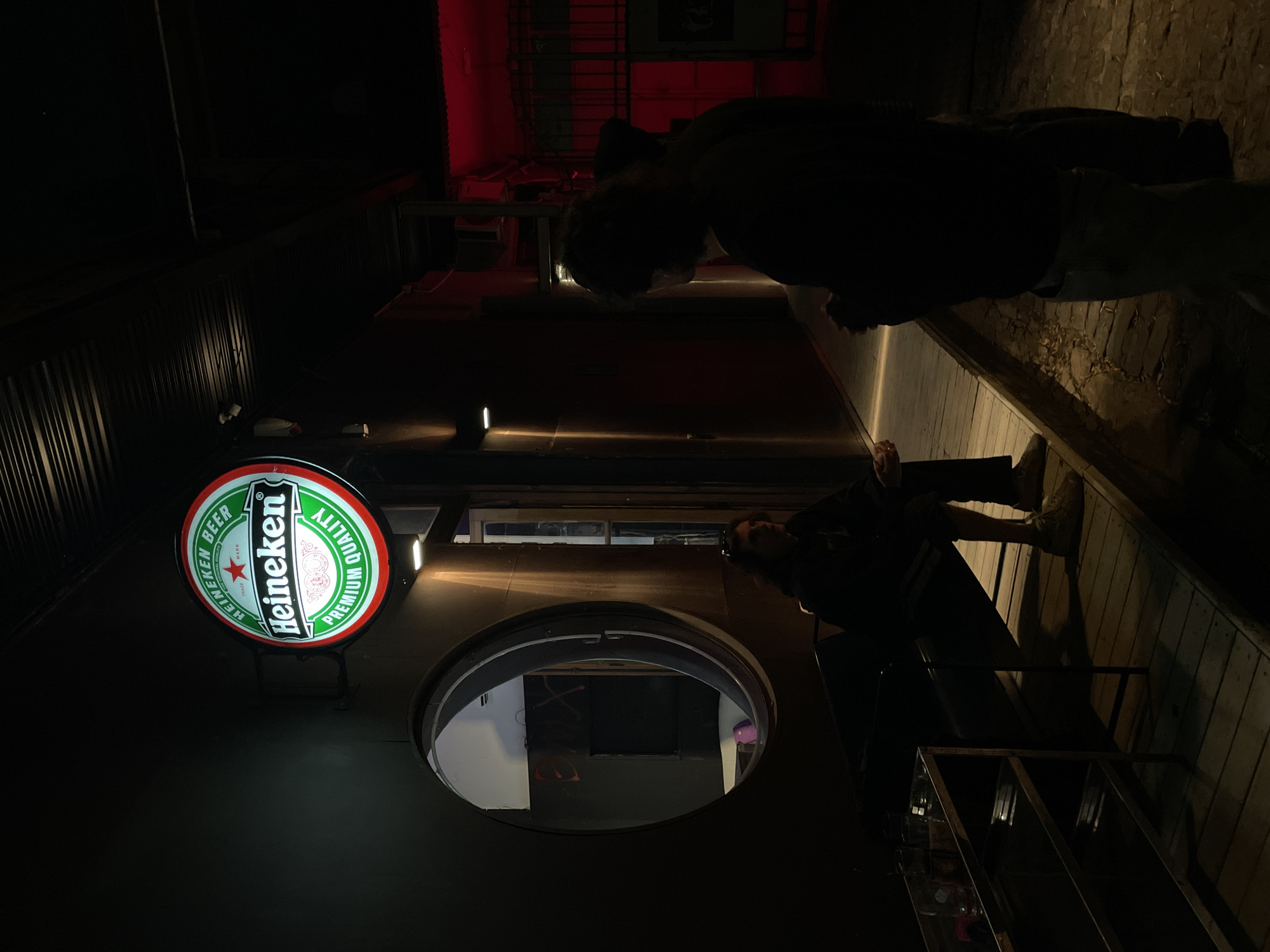 The image captures a nighttime urban scene, likely an alleyway or a street outside a establishment, consistent with locations found in Buenos Aires, Argentina. The overall lighting is dim, dominated by deep shadows and artificial light sources.
Two individuals are visible. One person, closer to the foreground on the right, is seen from the waist up, leaning against a dark surface or structure. They are mostly in shadow, but some light catches their face. Another figure is positioned further back on the left, partially obscured and bending forward, largely appearing as a silhouette against the dark background.
A prominent, illuminated circular Heineken beer sign is affixed to a dark wall on the left side of the frame. Below it, a large, dark circular window or opening reflects ambient light from within. To the right of the closer individual, a multi-tiered metal shelving unit is visible. The ground beneath the people consists of wooden planks, transitioning to what appears to be cobblestone further away. In the background, a strong red light emanates from an unseen source, casting a vibrant glow on a distant wall and structural elements.
The scene depicts a casual, still moment in a nighttime setting. The visible text on the illuminated sign reads: "Heineken BEER" and "PREMIUM QUALITY" around its edge, with "Heineken" in a larger font across the center and "MARK" below.
