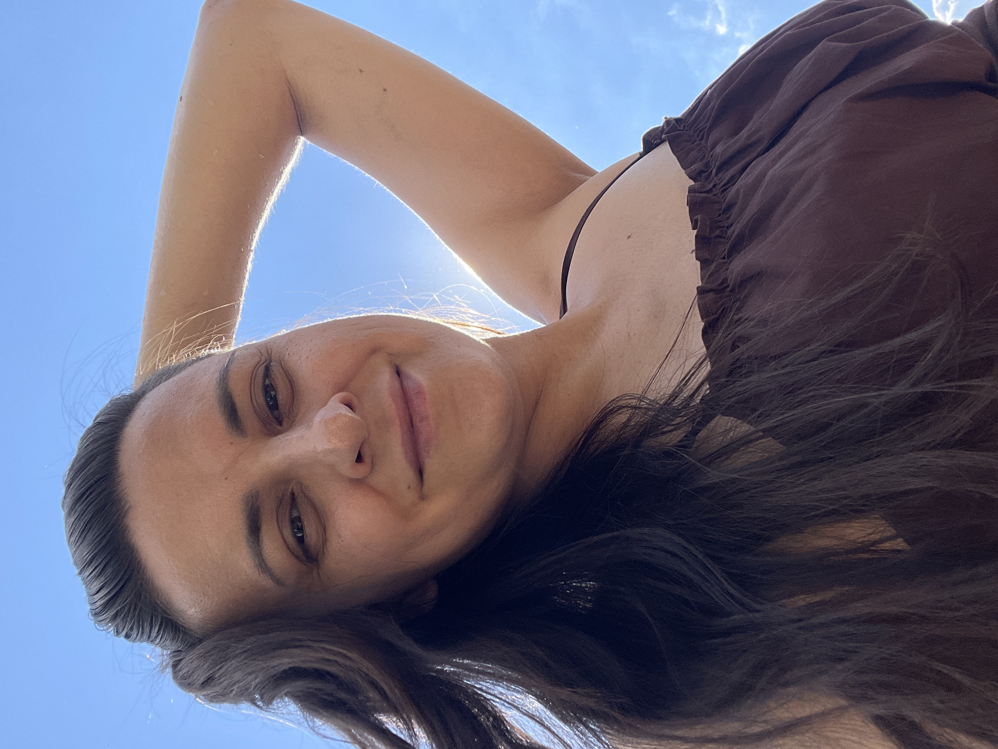 A low-angle, close-up, outdoor selfie shows a woman against a bright blue sky with scattered clouds. The woman has long, dark brown hair that is partially obscuring her face. She is looking directly at the camera with a slight smile. Her right arm is raised and bent, with her hand resting on her head. She is wearing a dark brown, strappy top. The sunlight is strong, creating a lens flare on the right side of the image, and backlighting her arm and hair. The scene appears to be in the city of Olkhovka, Russia, indicated by the bright, clear sky.