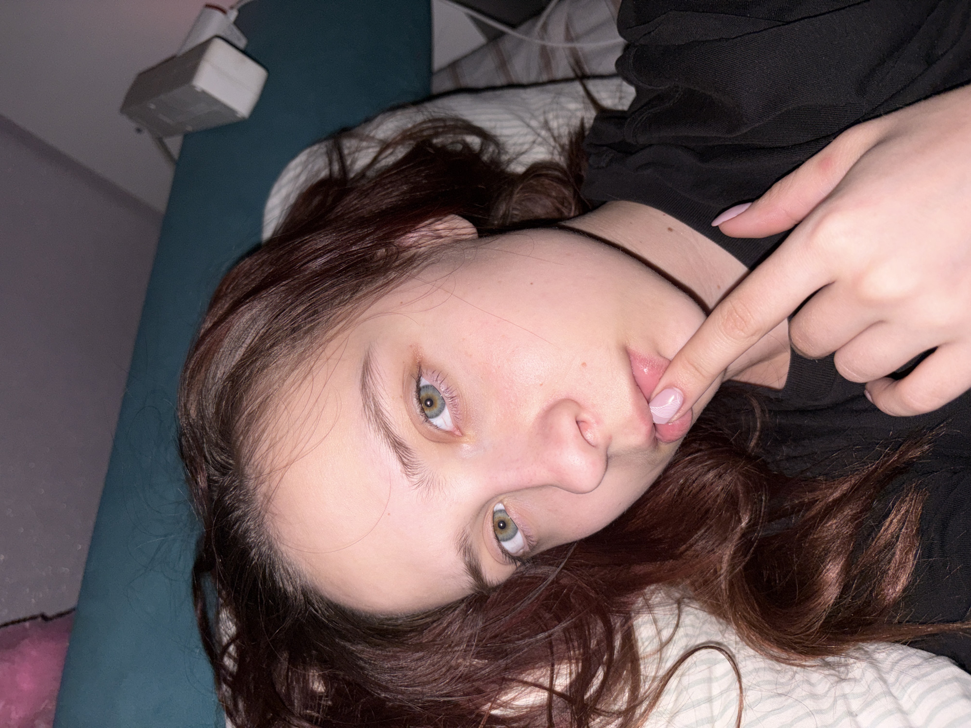 A young woman with brown hair and green eyes lies on her side in bed. She is wearing a black top. Her right index finger is touching her lower lip. The background includes a blue headboard and a white electrical outlet. The location is Прикамский, Russia.