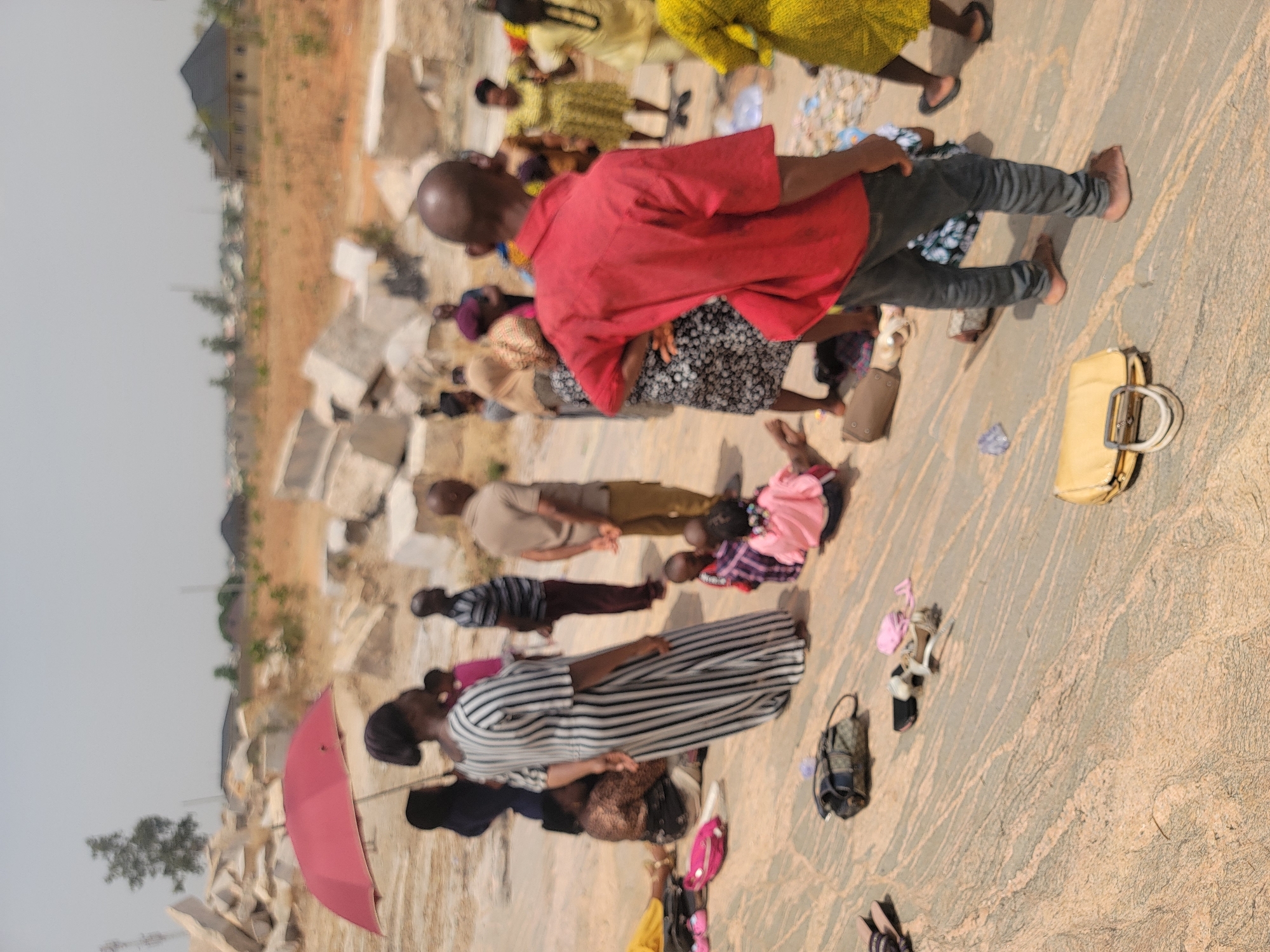 The image depicts a group of people in Narayi, Nigeria, some lying face down on what appears to be a stone surface, possibly praying or in a posture of submission. Individuals are dressed in diverse clothing, with examples of red, yellow, black-and-white striped patterns, and other casual attire. Several personal items are scattered around, including purses, shoes, and an umbrella. The surrounding environment includes construction materials, suggesting an incomplete building or a demolition site, and dry vegetation.