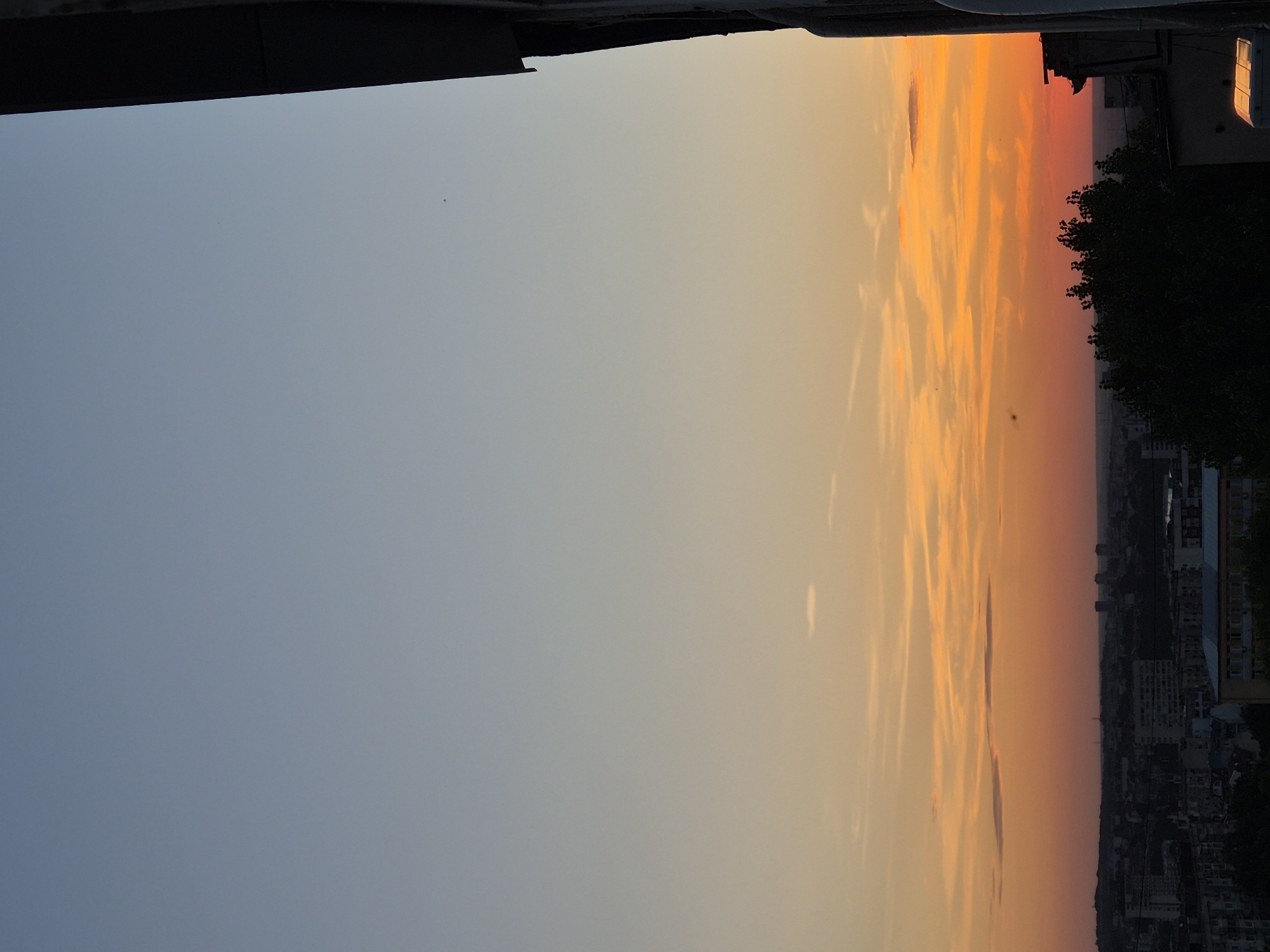 The image shows a sunset over a city. The sky is a gradient of blue at the top to orange and red at the horizon, with some scattered clouds. In the foreground, on the right side of the image, a dark silhouette of a building or structure is visible, partially obscuring the view of the city. The city itself is a dark mass of buildings with some lights visible. Trees are also visible in the foreground, silhouetted against the sky. The overall mood is serene and peaceful, with the vibrant colors of the sunset creating a dramatic backdrop. The specific location of the city is not identifiable from the image.
