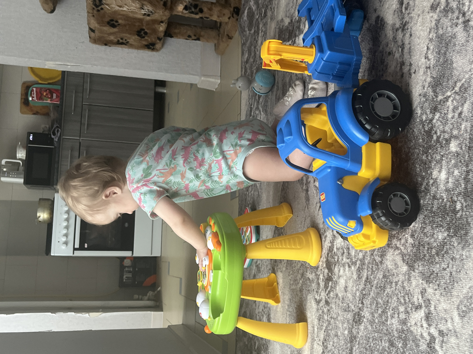 A child stands in a room that appears to be a kitchen. The child has blonde hair and wears a shirt with a dinosaur pattern. The child is interacting with a green and yellow toy table. A blue and yellow toy truck is nearby on a grey carpet. Behind the child is a kitchen stove and cabinets. A cat tree or similar pet structure is partially visible in the upper left corner. The location is Chaikovsky, Russia.