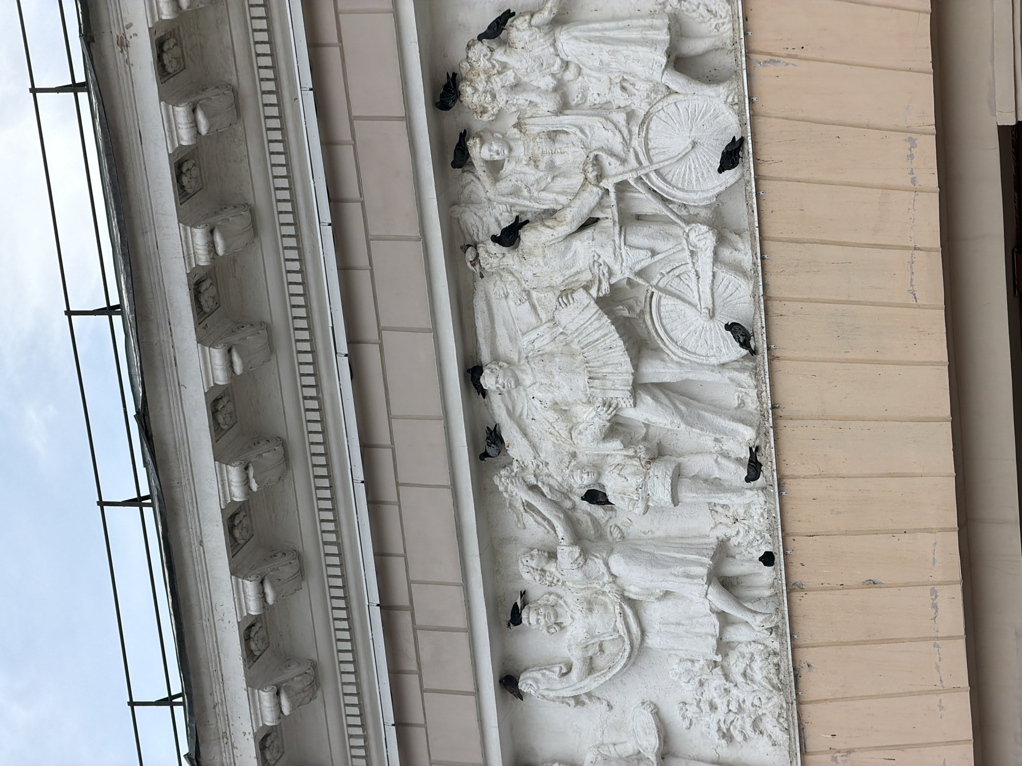 The image depicts a white stucco frieze on a building. The frieze features a bas-relief sculpture of several human figures, some of whom are interacting with a bicycle. There are multiple pigeons perched on and around the sculpture. Above the frieze, there is a white architectural molding with decorative elements. The building's facade above the molding is composed of light pink rectangular blocks. A dark railing with vertical supports is visible at the very top of the image, set against an overcast sky. The scene appears to be outdoors in an urban environment, likely in Moscow, Russia, given the architectural style which is common in Soviet-era and earlier Russian cities.